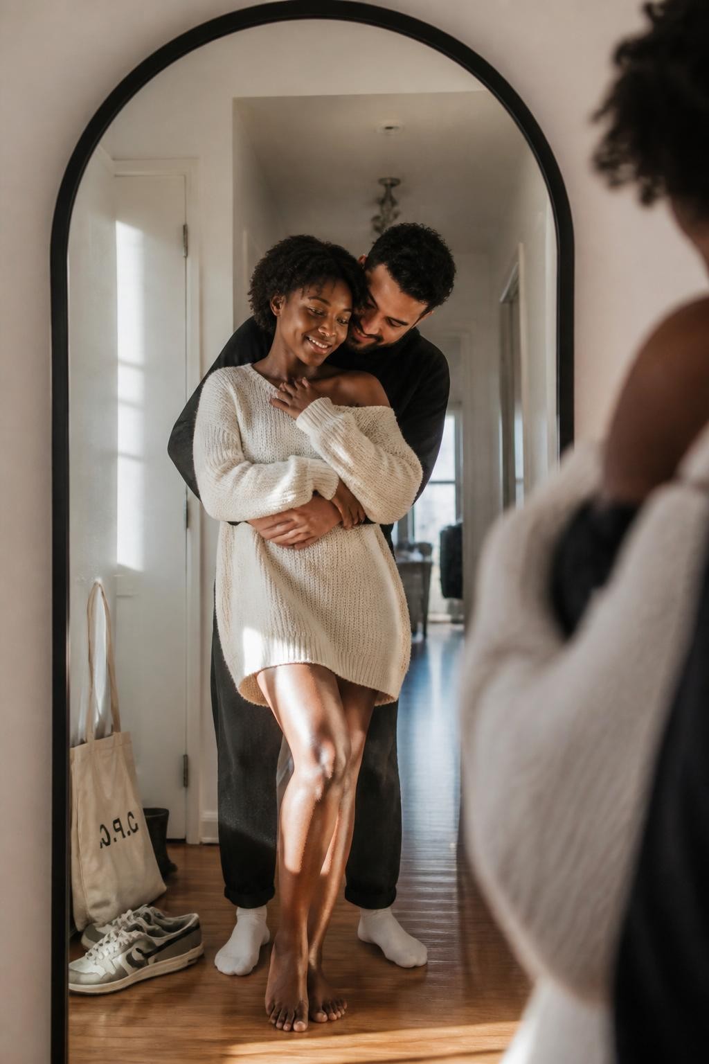 Image prompt: A modern romantic mirror portrait of an adult couple in a bright apartment hallway, reflected in a large arched black-framed mirror leaning against a white wall, featuring a Black woman with glowing dark skin and short natural curls wearing an oversized cream knit sweater, standing with her back partly toward the mirror while her partner hugs her gently from behind, both looking at their reflection with soft smiles, her hands resting over his arms, his chin near her shoulder without covering her face, warm afternoon light across the wooden floor, a canvas tote bag and sneakers near the doorway, slightly imperfect mirror streaks, relaxed home setting, professional lifestyle photography, natural pose, realistic texture, beautiful romantic feel.