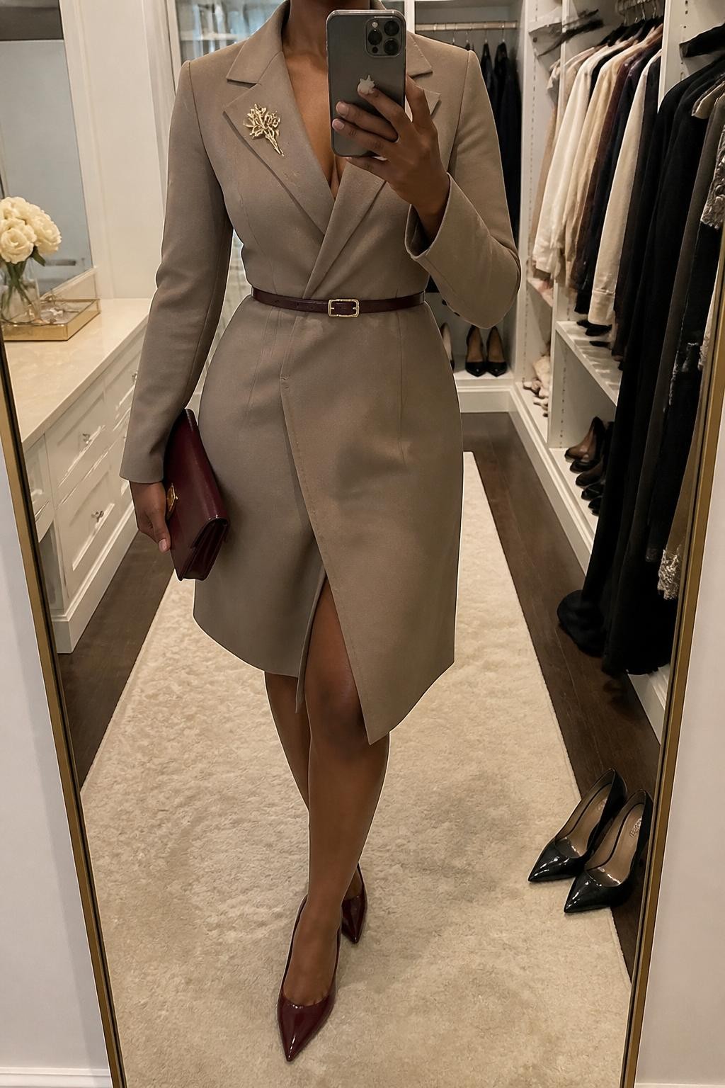 mirror selfie of a Black woman wearing the exact outfit described above, clearly showing a taupe coat dress with long sleeves, burgundy pointed pumps, a matching burgundy clutch, a thin brown waist belt, and a gold brooch on the lapel, holding a phone in front of a mirror, cropped at NECK so the face does not show, with a relaxed casual selfie pose, in a walk-in closet mirror, soft indoor lighting, with a pair of heels on the floor nearby and an open closet with clothes visible, latest iPhone photo quality.