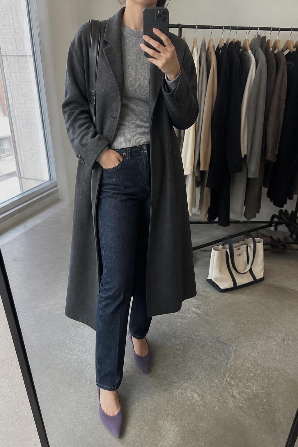 mirror selfie of a woman wearing the exact outfit described above, clearly showing a charcoal long coat layered over a soft gray fitted sweater, dark indigo straight-leg jeans, violet pointed flats, a black leather shoulder bag, and silver hoop earrings, holding a phone in front of a mirror, cropped at NECK so the face does not show, with a relaxed casual selfie pose, in a fashion studio mirror corner, natural window light, with clothing rack with garments behind and a tote bag resting on the floor, latest iPhone photo quality.