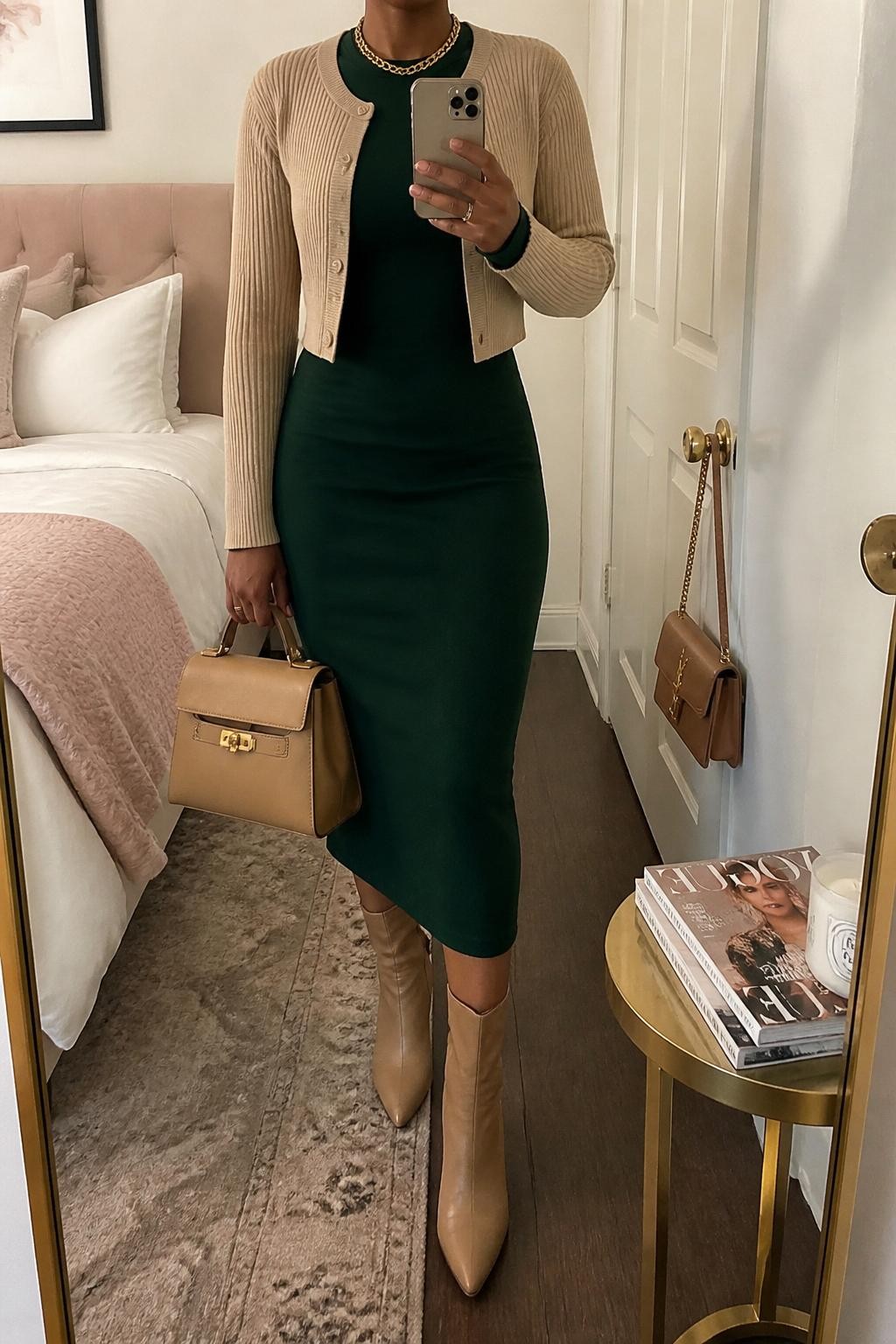 mirror selfie of a Black woman wearing the exact outfit described above, clearly showing a forest green long-sleeve midi dress, camel heeled boots, a beige cropped cardigan, a tan structured handbag, and a gold chain necklace, holding a phone in front of a mirror, cropped at NECK so the face does not show, with a relaxed casual selfie pose, in a feminine-themed bedroom, soft indoor lighting, with a handbag hanging on a door handle and a stack of fashion magazines on a side table, latest iPhone photo quality.