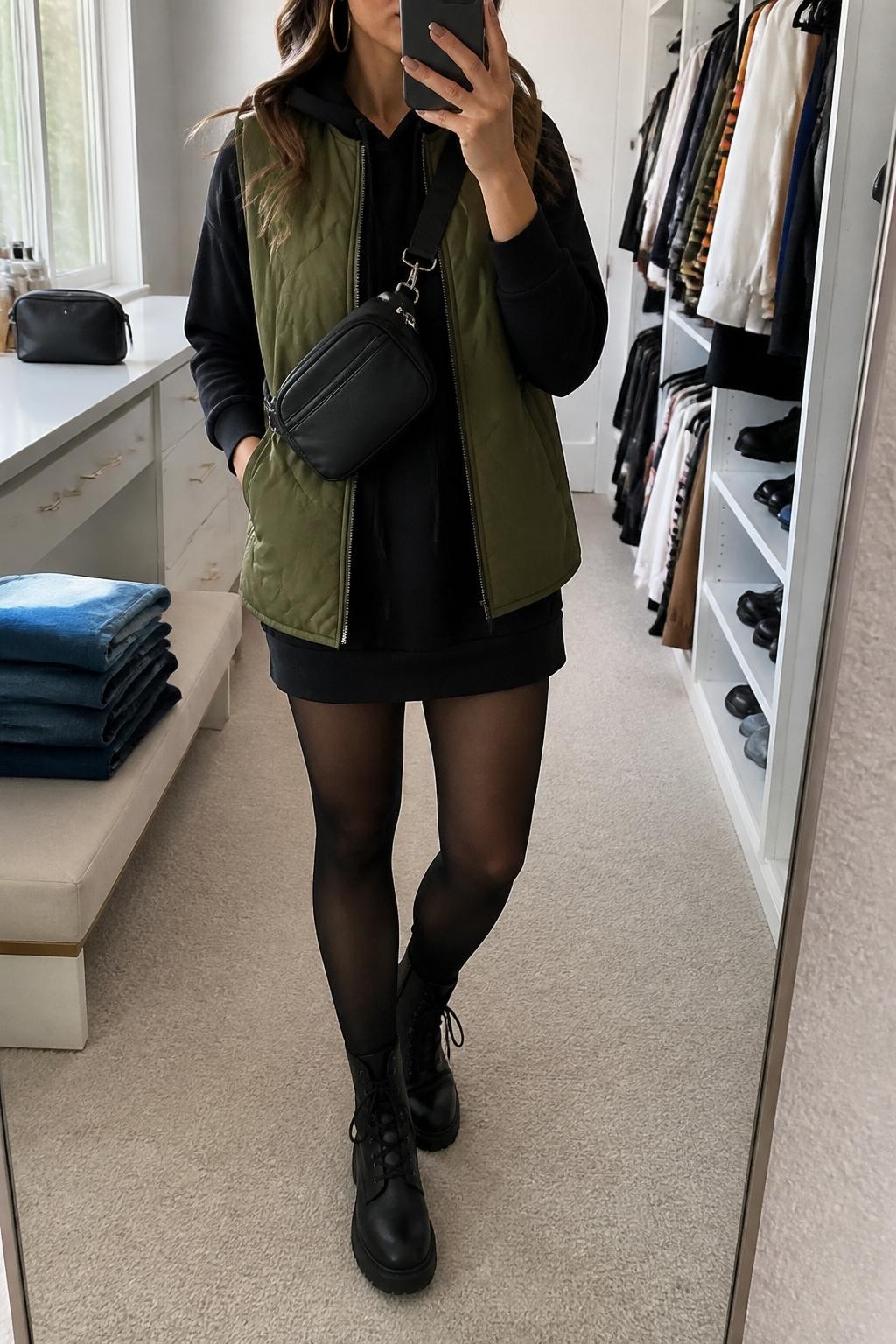 Mirror selfie of a woman wearing a black hoodie mini dress, olive green quilted vest, black opaque tights, black combat boots, gold hoop earrings, and a black nylon belt bag worn crossbody, holding a phone in front of a mirror, cropped at NECK so the face does not show, relaxed casual selfie pose, in a walk-in closet mirror, natural window light, with folded denim jeans on a bench and a cosmetic bag on the counter, latest iPhone photo quality.