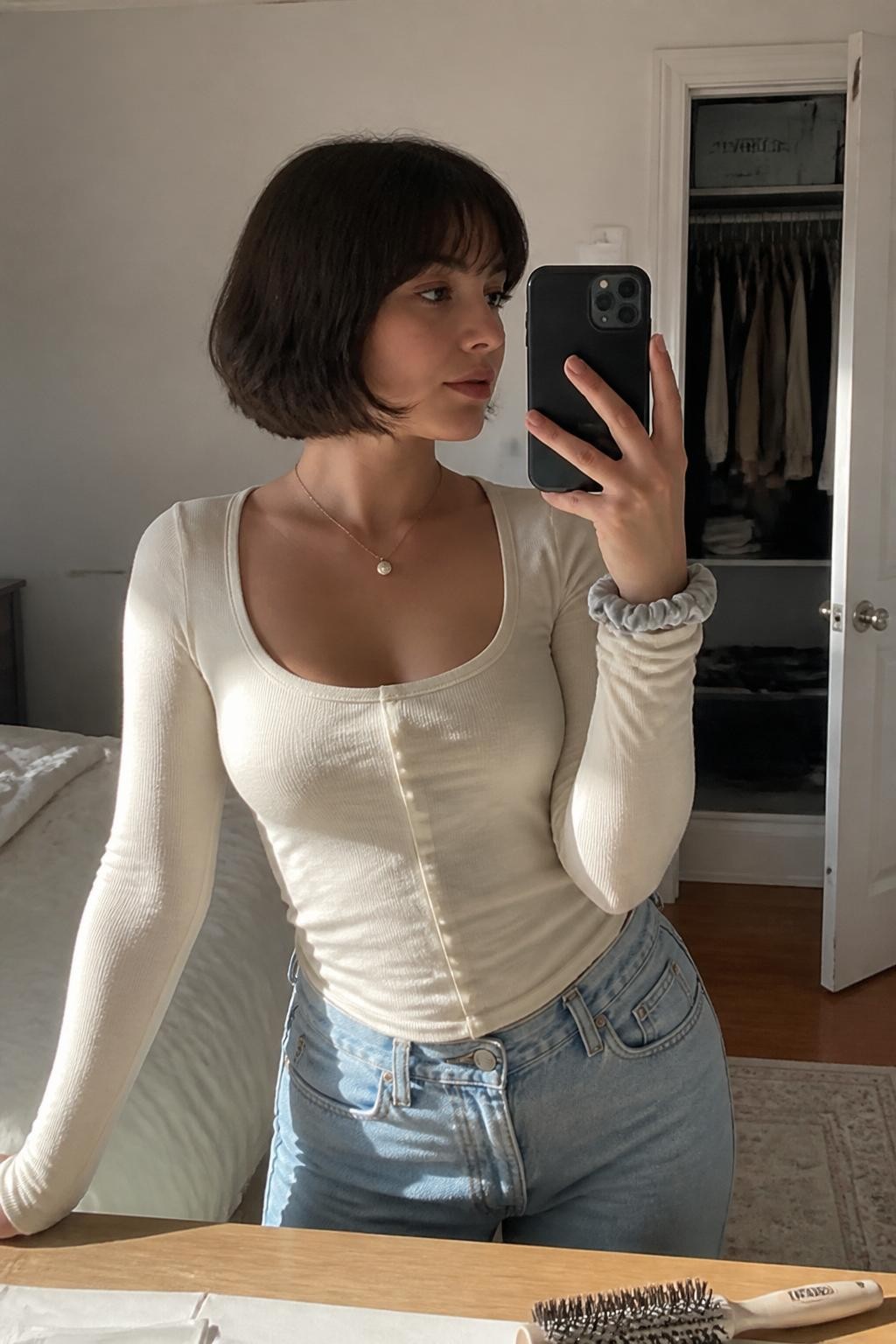 Image prompt: Photo of a woman wearing a chin-length rounded French bob with softly curved ends and light wispy bangs, candid mirror selfie, sun-drenched bedroom with a slightly open closet behind her, natural window light, containing a round brush on the dresser and a silk scrunchie around her wrist, realistic everyday getting-ready moment, latest iPhone photo quality.