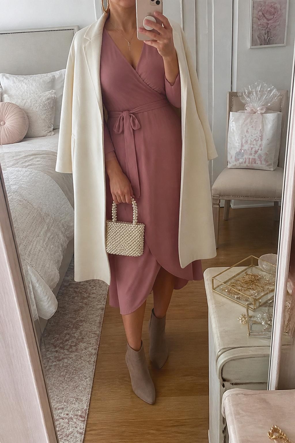 mirror selfie of a woman wearing the exact outfit described above, clearly showing a dusty rose long-sleeve wrap midi dress, taupe suede ankle boots, a cream wool coat draped open, a pearl mini handbag, and gold hoop earrings, holding a phone in front of a mirror, cropped at NECK so the face does not show, with a relaxed casual selfie pose, in a feminine bedroom mirror setup, soft natural window light, with a wrapped baby shower gift bag on a chair and a jewelry tray on the vanity, latest iPhone photo quality.