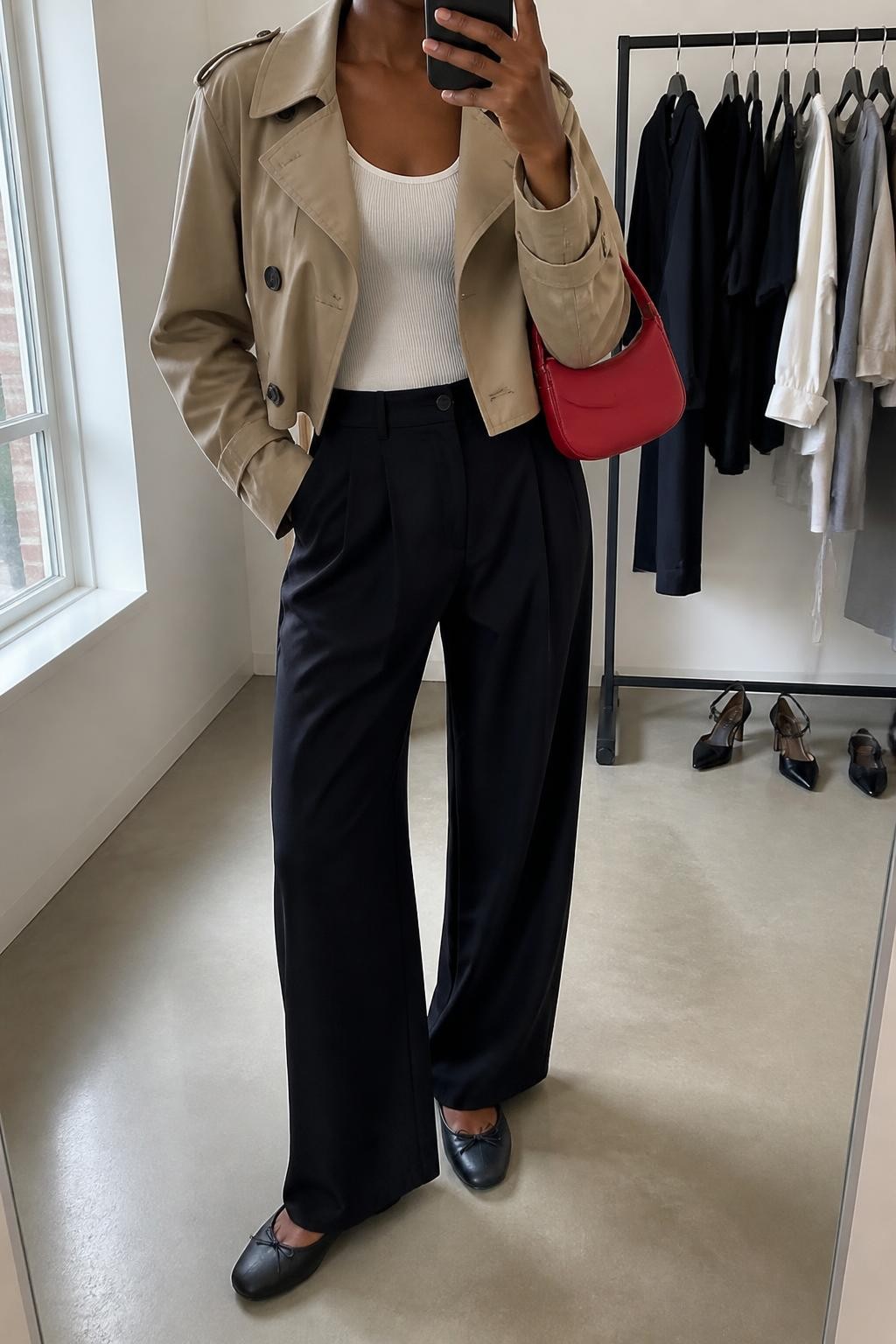 Mirror selfie of a Black woman wearing a cropped beige trench jacket over a fitted white ribbed tank top, black high-waisted wide-leg trousers in lightweight fabric, black leather ballet flats, and a vivid red mini shoulder bag, clearly showing the exact clothing items, colors, fabrics, and accessories listed, holding a phone in front of a minimal dressing room mirror, cropped at the neck so the face does not show, relaxed confident selfie pose, natural window light, a clothing rack with garments behind and a pair of heels on the floor nearby, polished and realistic, latest iPhone photo quality.