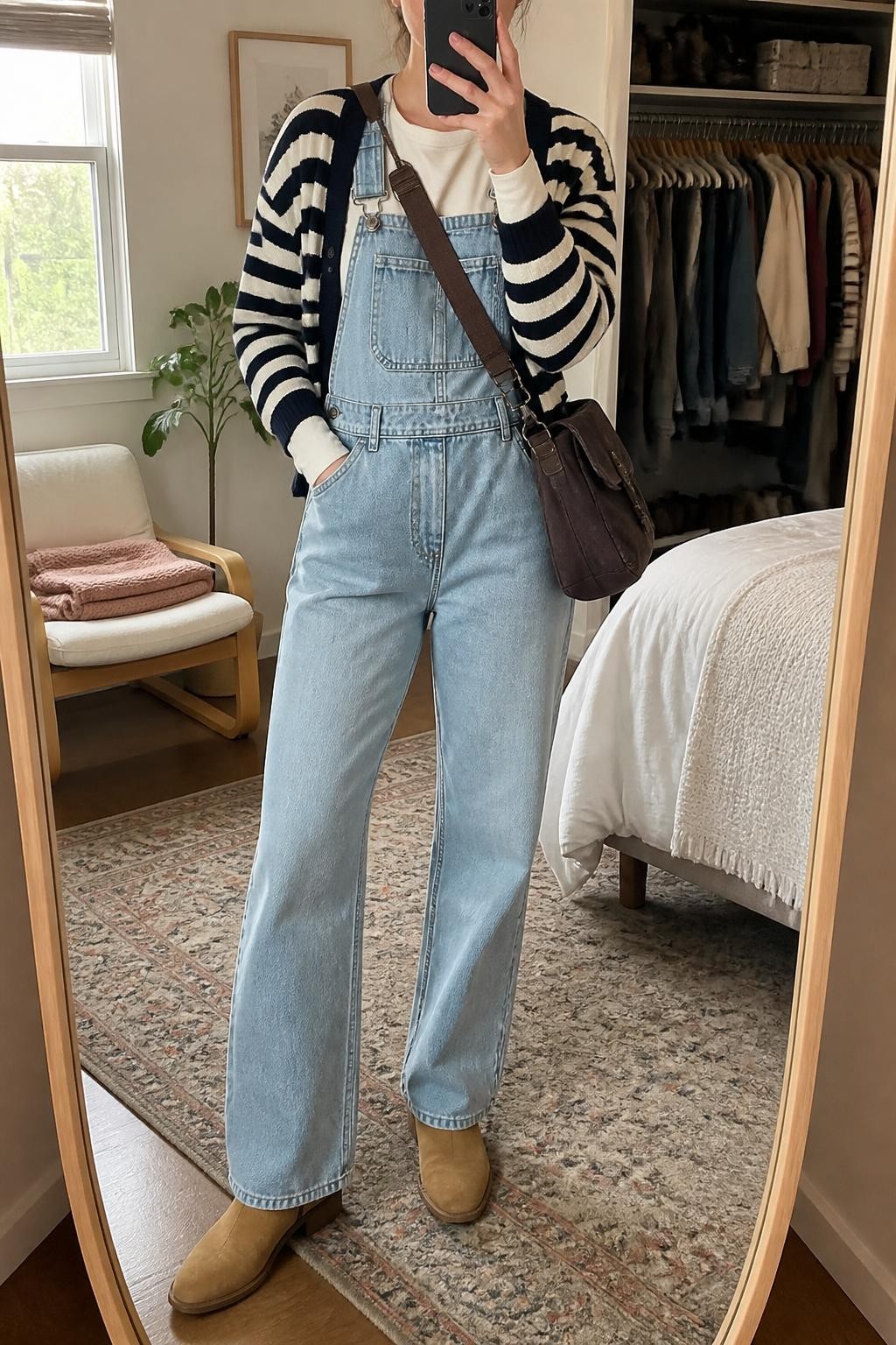 Mirror selfie of a woman wearing light-wash straight-leg denim overalls over a fitted cream long-sleeve top, a relaxed navy and ivory striped cardigan, tan suede ankle boots, and a dark brown canvas crossbody bag, clearly showing the exact clothing items, colors, fabrics, and accessories listed, holding a phone in front of a cozy bedroom mirror setup, cropped at the neck so the face does not show, relaxed casual selfie pose, natural window light, an open closet with clothes visible and a folded blanket on a chair, casual realistic setting, latest iPhone photo quality.