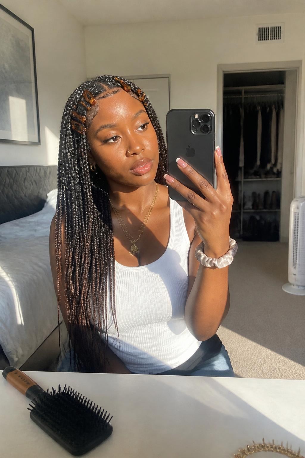 Image prompt: Photo of a Black woman wearing long dark brown knotless braids decorated with several small tortoiseshell clips near the front, candid mirror selfie, sun-drenched bedroom with a slightly open closet behind her, natural window light, containing a hairbrush on the dresser and a silk scrunchie around her wrist, realistic everyday getting-ready moment, latest iPhone photo quality.