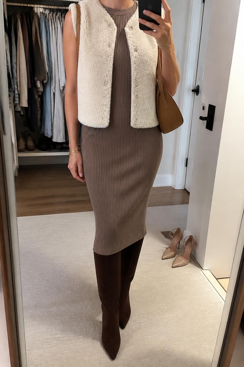 Mirror selfie of a woman wearing a cream faux-shearling vest over a fitted mocha ribbed-knit midi dress, chocolate-brown suede knee-high boots, a structured tan shoulder bag, and thin gold bangles, clearly showing the exact clothing items, colors, fabrics, and accessories listed, holding a phone in front of a walk-in closet mirror, cropped at the neck so the face does not show, relaxed casual selfie pose, soft indoor lighting, an open closet with clothes visible and a pair of heels on the floor nearby, cozy and uncluttered, latest iPhone photo quality.