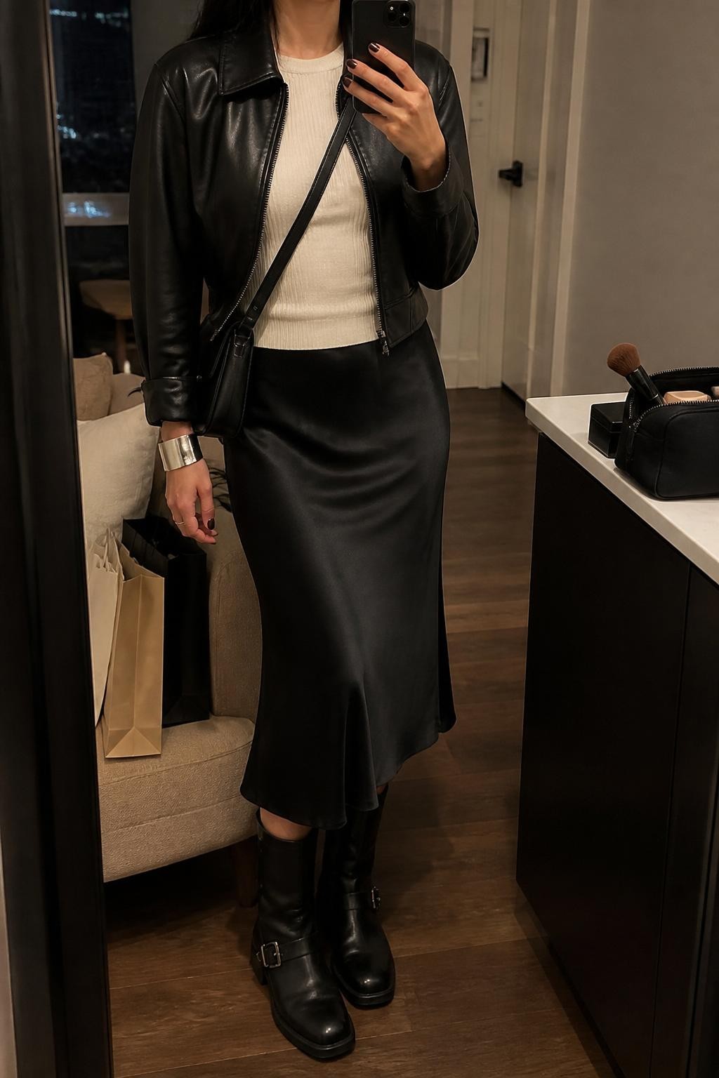 mirror selfie of a woman wearing the exact outfit described above, clearly showing black moto boots with subtle buckle details, a black satin midi skirt, an ivory fitted ribbed top, a cropped black leather jacket, a black crossbody bag, and silver cuff bracelet, holding a phone in front of a mirror, cropped at NECK so the face does not show, with a relaxed casual selfie pose, in a stylish city apartment, warm moody lighting, with shopping bags beside a chair and a makeup brush or cosmetic bag on the counter, latest iPhone photo quality.