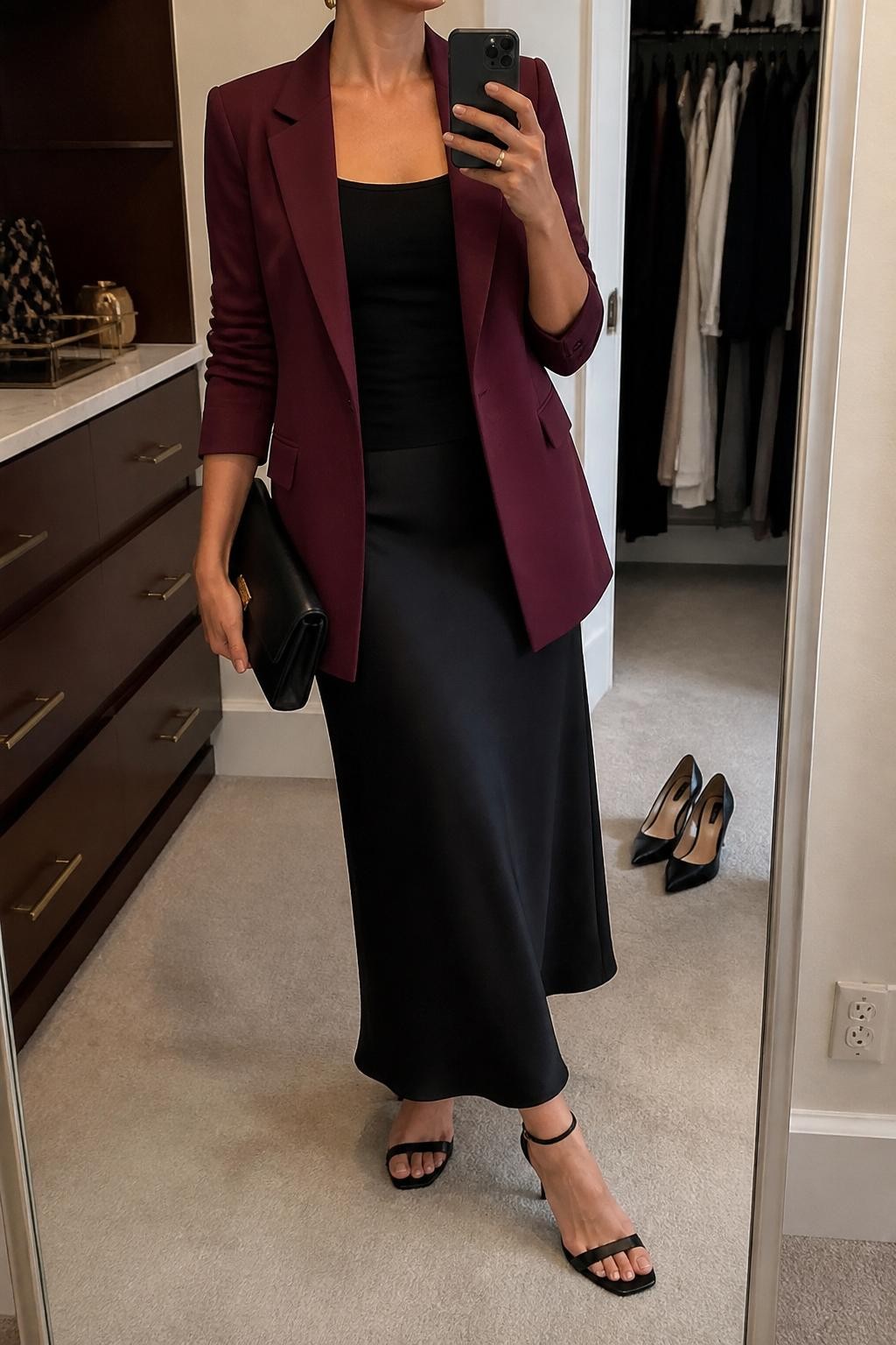 mirror selfie of a woman wearing the exact outfit described above, clearly showing a wine red tailored blazer layered over a fitted black tank, black satin maxi skirt, black ankle-strap heels, a black envelope clutch, and gold stud earrings, holding a phone in front of a mirror, cropped at NECK so the face does not show, with a relaxed casual selfie pose, in a walk-in closet mirror, soft indoor lighting, with a pair of heels on the floor nearby and an open closet with clothes visible, latest iPhone photo quality.