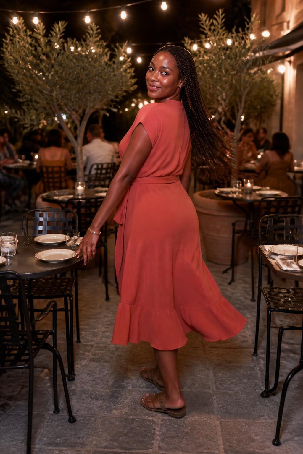 Image prompt: A romantic outdoor restaurant patio photo of an adult Black woman with deep brown skin standing between small patio tables under string lights, wearing a soft terracotta wrap dress and flat sandals, turning back toward the camera while lightly touching the back of a metal chair, her braids moving gently in the evening breeze, small plates and water glasses on nearby tables, potted olive trees, warm string lights overhead, stone floor, realistic fabric movement, natural smile, DSLR patio dinner photography with a warm modern feel.