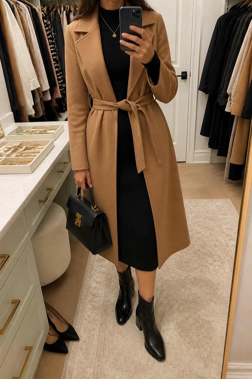 mirror selfie of a woman wearing the exact outfit described above, clearly showing a camel wrap coat layered over a black knit midi dress with long sleeves, black block heel ankle boots, a structured black handbag, and a slim gold pendant necklace, holding a phone in front of a mirror, cropped at NECK so the face does not show, with a relaxed casual selfie pose, in a walk-in closet mirror, soft indoor lighting, with a jewelry tray on the vanity and a pair of heels on the floor nearby, latest iPhone photo quality.