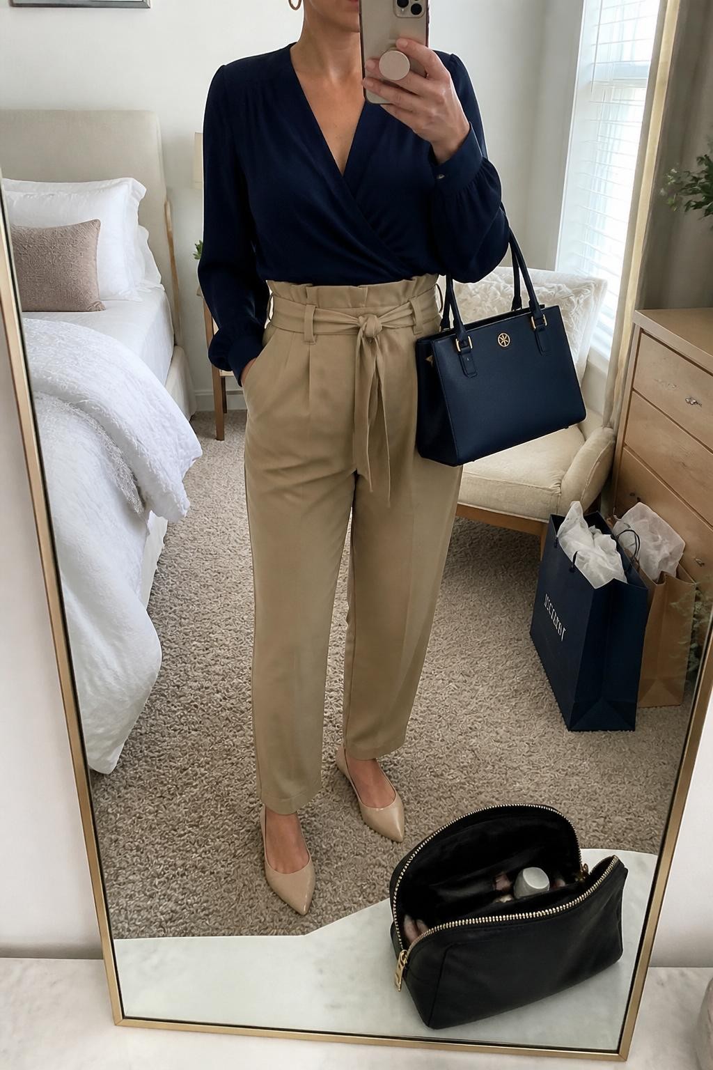 Mirror selfie of a woman wearing a navy wrap blouse, tan paperbag waist trousers, nude pointed flats, gold hoop earrings, and a navy structured handbag, cropped at neck so the face does not show, in a cozy bedroom mirror setup with natural window light, a cosmetic bag on the counter and shopping bags beside a chair, realistic latest iPhone photo quality.