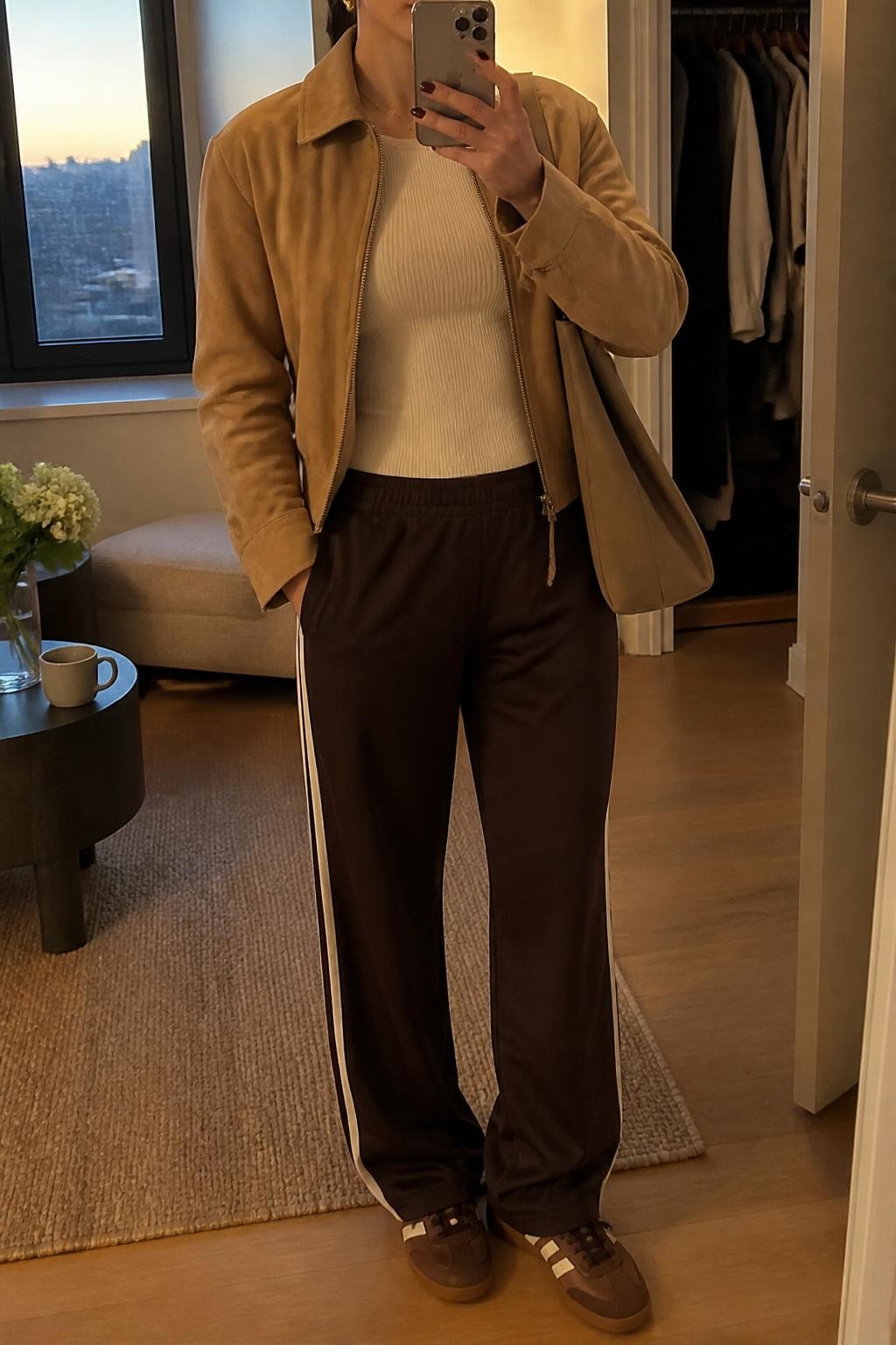 Mirror selfie of a woman wearing a tan suede jacket, cream fitted ribbed top, dark brown track pants with cream side stripes, brown retro sneakers, gold stud earrings, and a tan canvas tote bag, holding a phone in front of a mirror, cropped at NECK so the face does not show, relaxed casual selfie pose, in a stylish city apartment, evening golden hour, with a coffee mug on a side table and an open closet with clothes visible, latest iPhone photo quality.