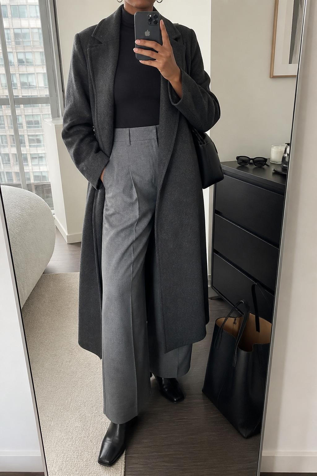 Mirror selfie of a Black woman wearing a long charcoal wool coat left open over a fitted black mock-neck top and heather gray wide-leg tailored trousers, clearly showing black leather square-toe ankle boots, a structured black shoulder bag, and small silver hoop earrings, holding an iPhone in front of a stylish city apartment mirror, cropped at the neck so the face does not show, relaxed casual selfie pose, natural window light, sunglasses placed on a dresser and a tote bag resting on the floor, natural unstaged setting, latest iPhone photo quality.