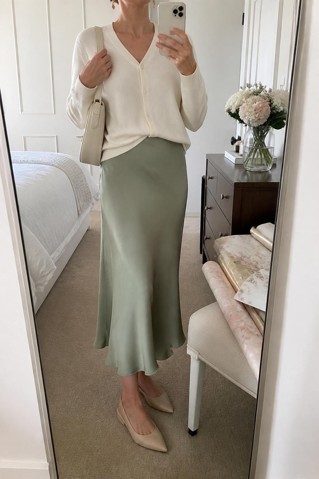 mirror selfie of a woman wearing the exact outfit described above, clearly showing a sage green satin midi slip skirt, ivory button-front cardigan worn softly tucked, nude slingback flats, a cream shoulder bag, and delicate gold drop earrings, holding a phone in front of a mirror, cropped at NECK so the face does not show, with a relaxed casual selfie pose, in a bright bedroom mirror setup, soft morning light, with pastel gift wrapping paper on a chair and a small bouquet on the dresser, latest iPhone photo quality.