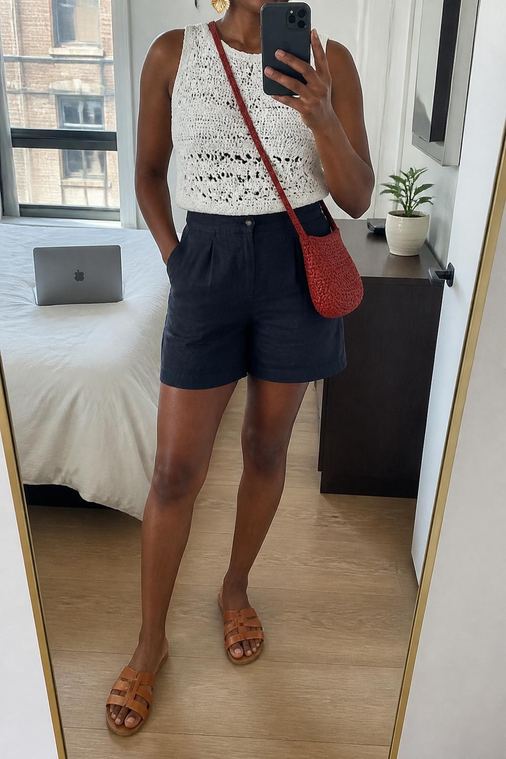 mirror selfie of a Black woman wearing a white sleeveless crochet top, navy high-waisted linen shorts, tan leather slide sandals, red woven crossbody bag, and gold shell earrings, clearly showing the same clothing items, colors, fabrics, and accessories listed in the Outfit Pieces section, holding a phone in front of a mirror, cropped at NECK so the face does not show, with a relaxed casual selfie pose, in a stylish city apartment, natural window light, including a laptop on the bed and a small plant on a dresser, latest iPhone photo quality.