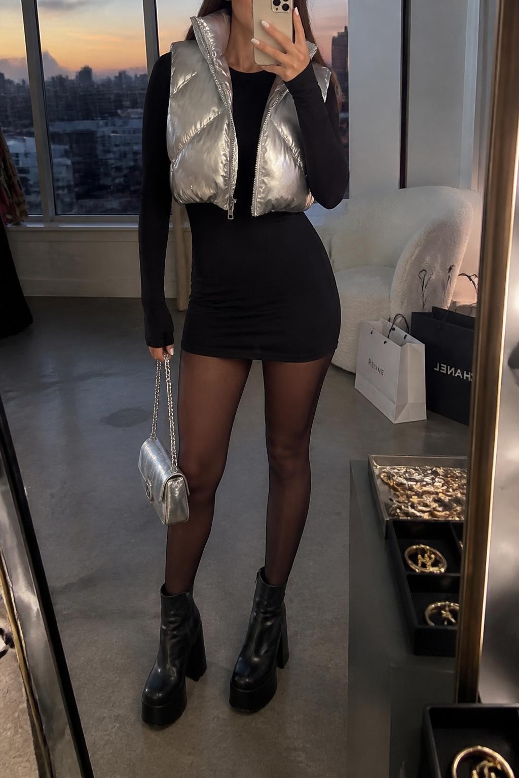 mirror selfie of a woman wearing a silver cropped puffer vest over a black long-sleeve mini dress, sheer black tights, black platform ankle boots, and a silver metallic mini bag, clearly showing the same clothing items, colors, fabrics, and accessories listed in the Outfit Pieces section, holding a phone in front of a mirror, cropped at NECK so the face does not show, with a relaxed casual selfie pose, in a fashion studio mirror corner, evening golden hour, including shopping bags beside a chair and a jewelry tray on the vanity, latest iPhone photo quality.