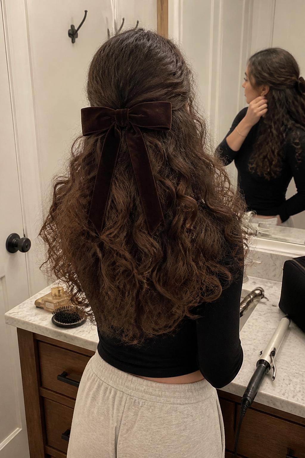 Image prompt: Photo of a woman wearing a full brushed-out curly ponytail with a soft cloud-like texture and a dark brown velvet bow, back-of-head view to highlight the shape, bathroom vanity mirror with a hairbrush and perfume bottle nearby, soft indoor lighting, containing a curling iron on the counter and a makeup bag beside the sink, realistic everyday moment, latest iPhone photo quality.