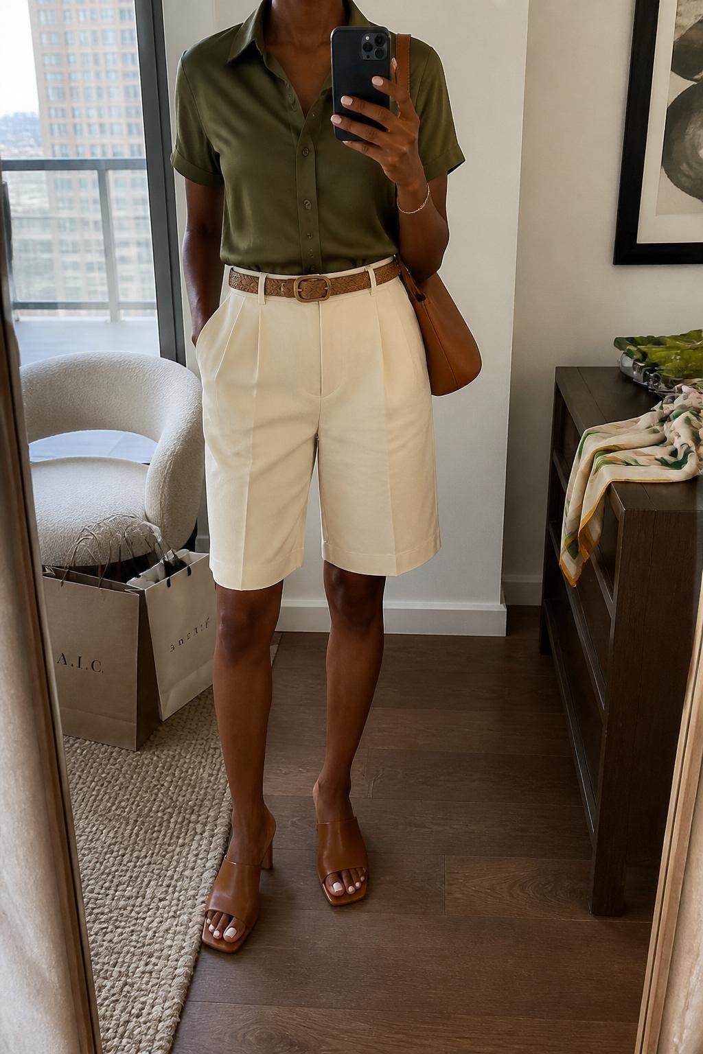 mirror selfie of a Black woman wearing the exact outfit described above, clearly showing cream tailored knee-length shorts, olive silk short-sleeve blouse tucked in, tan woven belt, brown leather mules, a caramel shoulder bag, and pearl stud earrings, holding a phone in front of a mirror, cropped at NECK so the face does not show, with a relaxed casual selfie pose, in a stylish city apartment mirror, warm indoor light, with shopping bags beside a chair and a folded scarf on a dresser, latest iPhone photo quality.