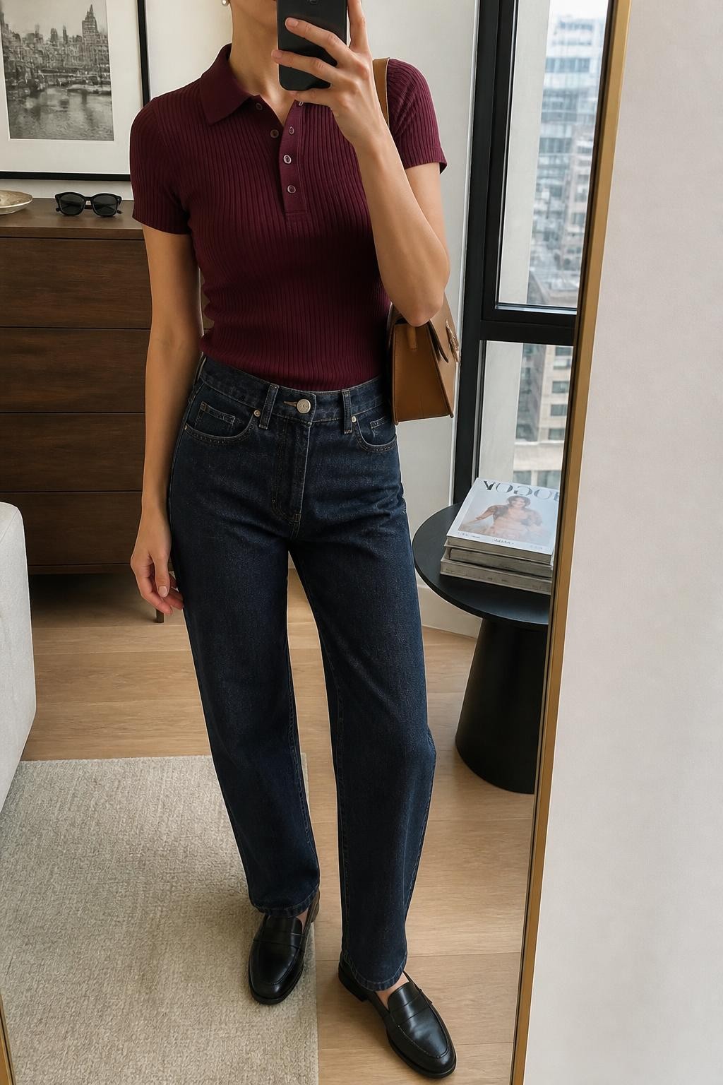 Mirror selfie of a woman wearing a fitted burgundy ribbed short-sleeve polo shirt, dark indigo relaxed straight-leg jeans, black leather loafers, structured camel shoulder bag, and small pearl stud earrings, clearly showing the exact clothing items, colors, fabrics, and accessories listed, holding a phone in front of a stylish city apartment mirror, cropped at the neck so the face does not show, relaxed casual selfie pose, soft indoor lighting, sunglasses placed on a dresser and a stack of fashion magazines on a side table, natural and uncluttered, latest iPhone photo quality.