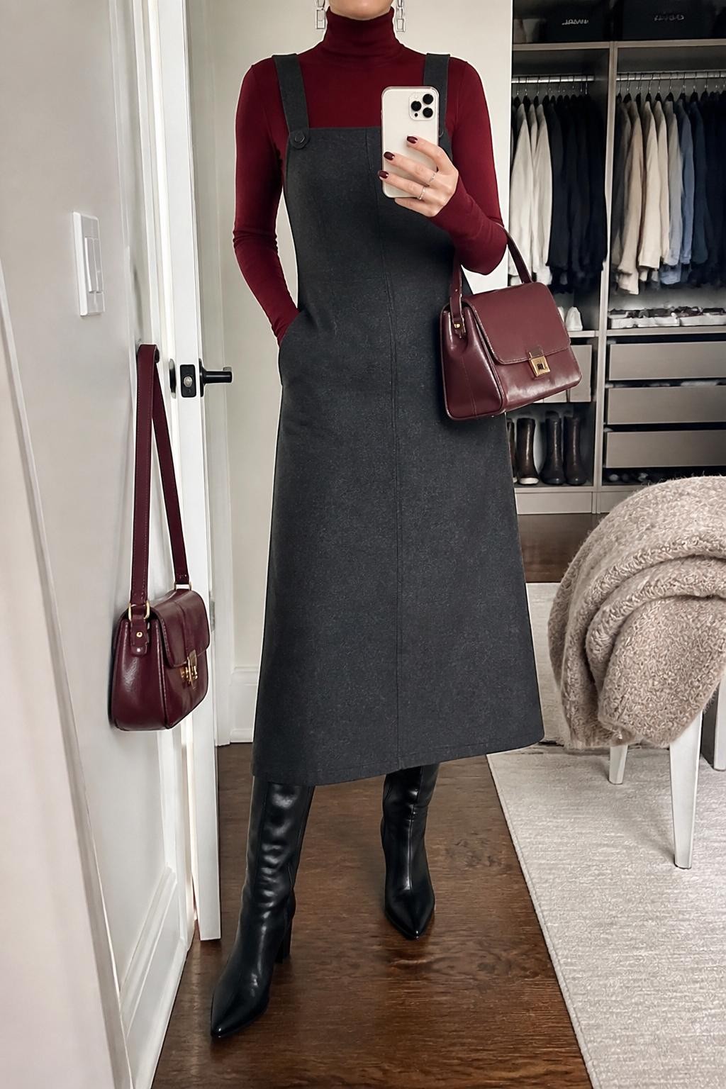 Mirror selfie of a woman wearing a charcoal gray wool-blend overall midi dress over a fitted burgundy turtleneck, clearly showing black knee-high leather boots, a structured burgundy handbag, and silver geometric earrings, holding an iPhone in front of a walk-in closet mirror, cropped at the neck so the face does not show, relaxed casual selfie pose, soft indoor lighting, a handbag hanging on a door handle and a folded blanket on a chair, natural unstaged setting, latest iPhone photo quality.
