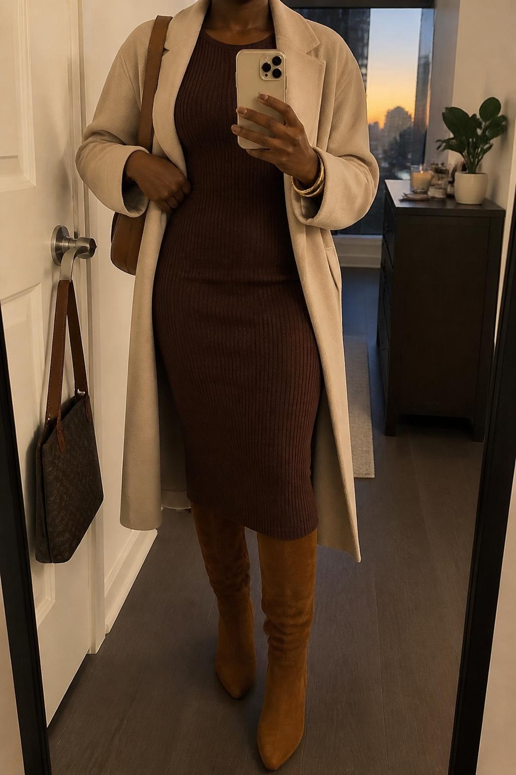 mirror selfie of a Black woman wearing the exact outfit described above, clearly showing a chocolate brown ribbed knit midi dress, caramel suede knee-high boots, a beige belted coat, a caramel shoulder bag, and gold bangles, holding a phone in front of a mirror, cropped at NECK so the face does not show, with a relaxed casual selfie pose, in a stylish city apartment, evening golden hour, with a handbag hanging on a door handle and a small plant on a dresser, latest iPhone photo quality.