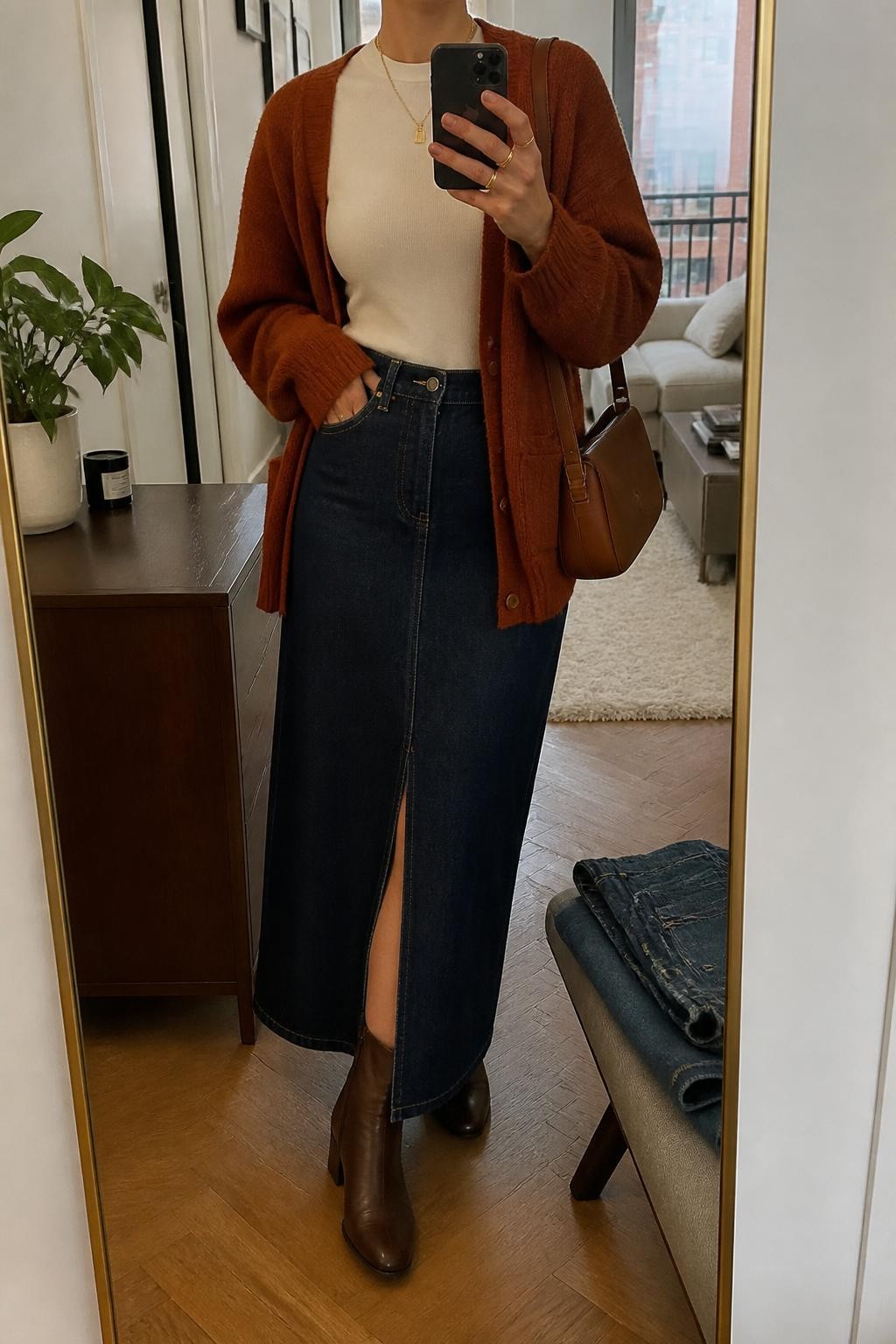 Mirror selfie of a woman wearing a dark indigo denim maxi skirt with a front slit, a fitted cream crew-neck top, an oversized rust-orange button-front cardigan, chocolate-brown leather ankle boots, and a cognac shoulder bag, clearly showing the exact clothing items, colors, fabrics, and accessories listed, holding a phone in front of a stylish city apartment mirror, cropped at the neck so the face does not show, relaxed casual selfie pose, warm moody lighting, a small plant on a dresser and folded denim jeans on a bench, believable everyday scene, latest iPhone photo quality.