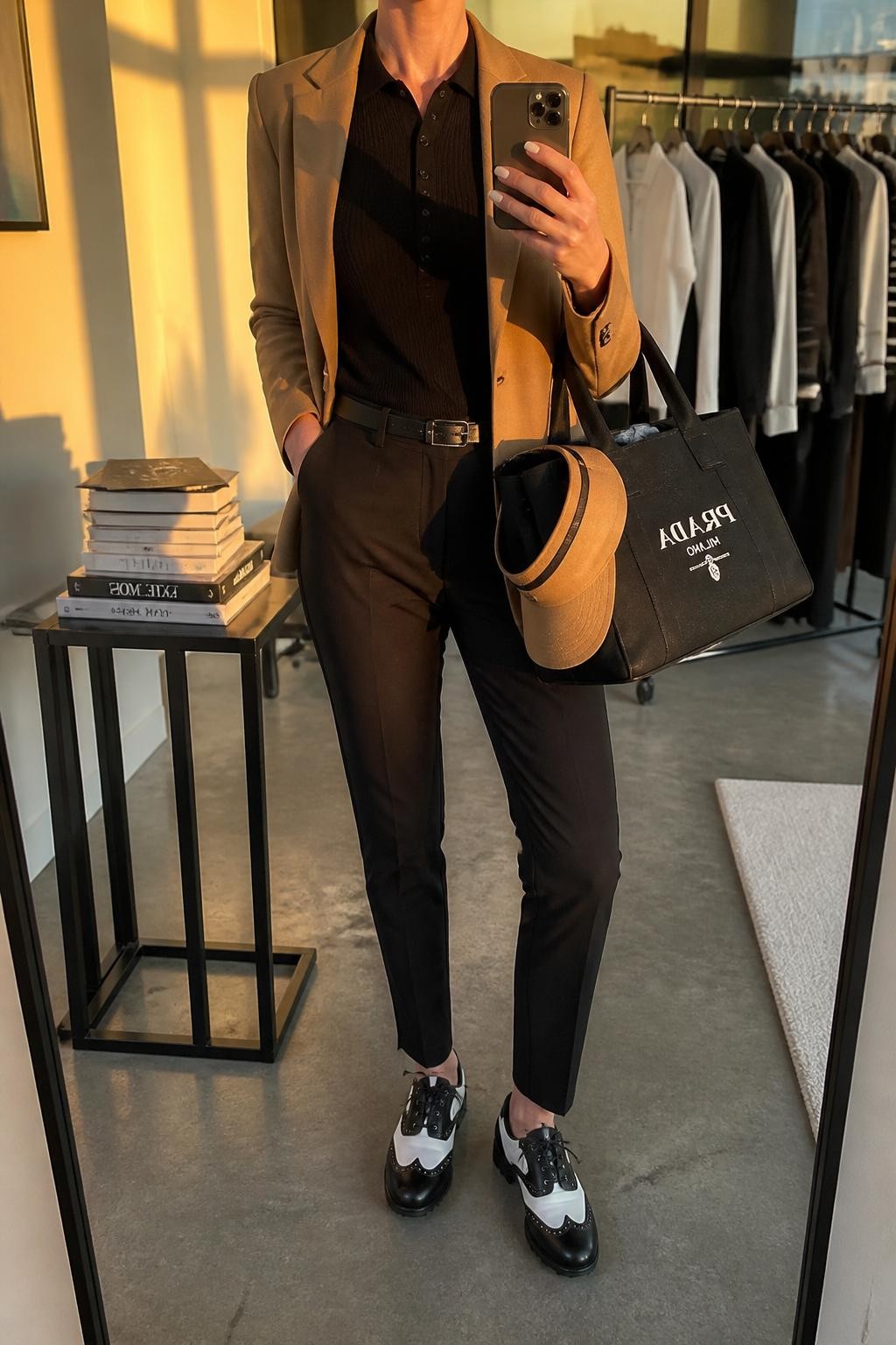 Mirror selfie of a woman wearing a camel unstructured stretch golf blazer over a fitted black knit polo, paired with black tapered golf trousers, clearly showing black-and-white leather golf shoes, a camel visor, and a structured black golf tote, holding an iPhone in front of a fashion studio mirror corner, cropped at the neck so the face does not show, relaxed casual selfie pose, evening golden hour light, a stack of fashion magazines on a side table and a clothing rack with garments behind, natural unstaged setting, latest iPhone photo quality.