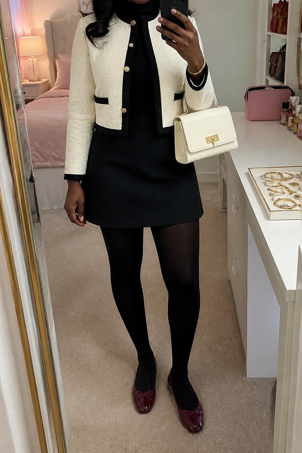 Mirror selfie of a Black woman wearing a cropped ivory bouclé jacket with black trim, fitted black turtleneck, black A-line mini skirt, opaque black tights, burgundy leather ballet flats, and a structured ivory handbag, clearly showing the exact clothing items, colors, fabrics, and accessories listed, holding a phone in front of a feminine-themed bedroom mirror, cropped at the neck so the face does not show, relaxed casual selfie pose, soft indoor lighting, a jewelry tray on the vanity and a makeup bag on the counter, polished yet natural, latest iPhone photo quality.