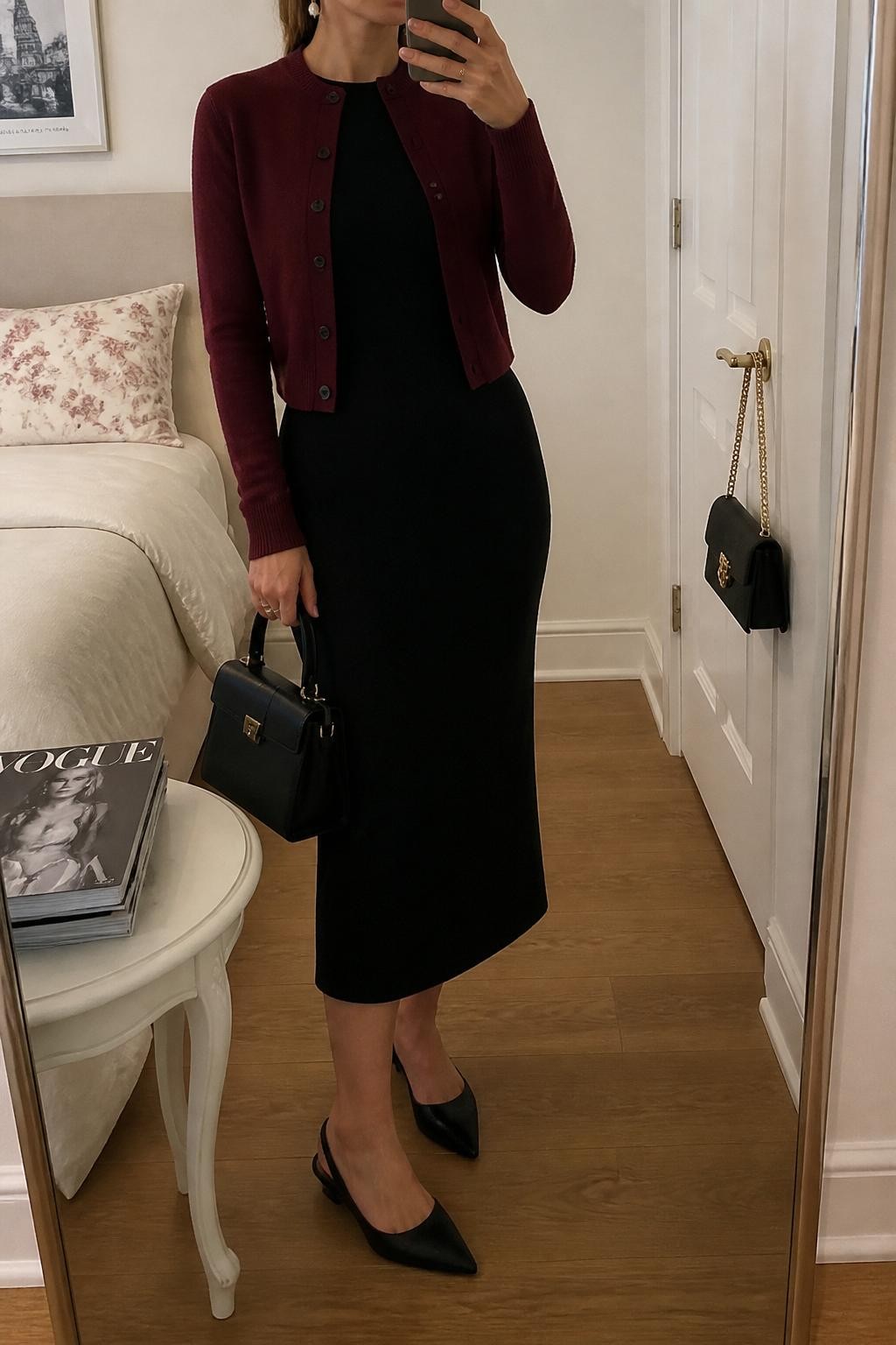 Mirror selfie of a woman wearing a black tailored midi dress, burgundy button-front cardigan, black slingback flats, pearl drop earrings, and a small black top-handle bag, cropped at neck so the face does not show, in a feminine-themed bedroom with soft indoor lighting, a handbag hanging on a door handle and a stack of fashion magazines on a side table, realistic latest iPhone photo quality.