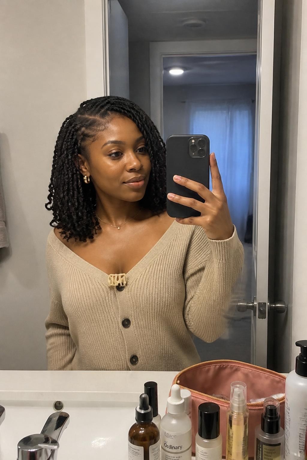 Image prompt: Photo of a Black woman wearing shoulder-length passion twists with soft loose ends and a natural side part, candid mirror selfie, bathroom vanity mirror with skincare bottles and a makeup bag nearby, soft indoor lighting, containing a claw clip clipped to the neckline of her cardigan, realistic everyday getting-ready moment, latest iPhone photo quality.