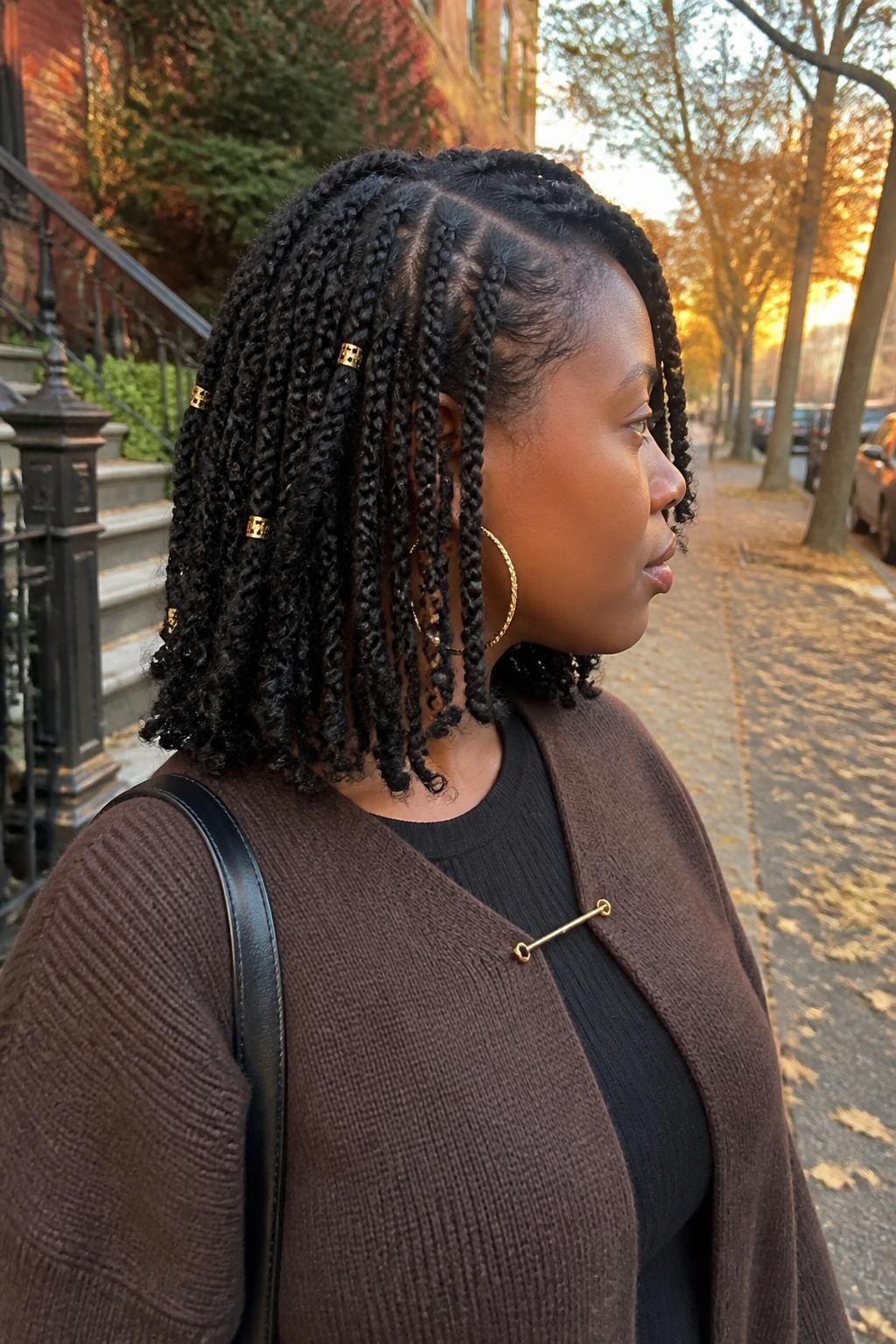 Image prompt: Photo of a Black woman wearing a shoulder-length mini twist bob with a deep side part and a few small gold cuffs, side profile view, softly blurred city street background with scattered autumn leaves, golden hour glow, containing hoop earrings peeking through and a slim hair clip attached to her cardigan collar, realistic candid photo, latest iPhone photo quality.