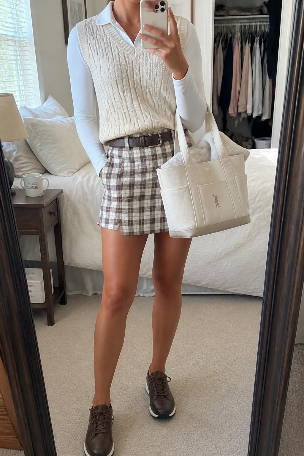 Mirror selfie of a woman wearing a cream cable-knit sweater vest over a white fitted long-sleeve collared golf shirt, paired with a brown-and-cream plaid golf skort with built-in shorts, clearly showing chocolate brown spikeless golf shoes, a dark brown leather belt, and a structured cream golf tote, holding an iPhone in front of a cozy bedroom mirror setup, cropped at the neck so the face does not show, relaxed casual selfie pose, natural window light, a coffee mug on a bedside table and an open closet with clothes visible, natural unstaged setting, latest iPhone photo quality.