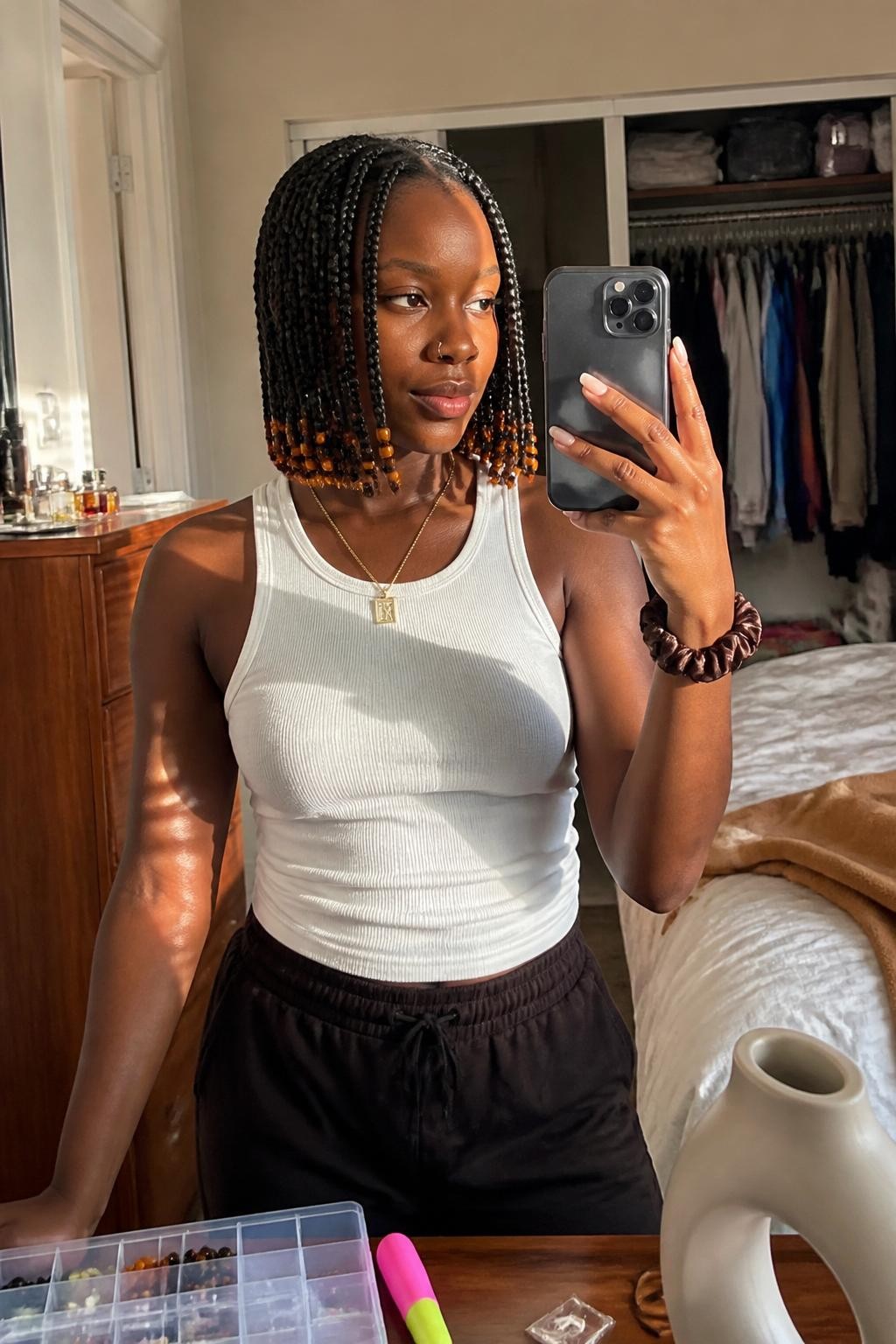 Image prompt: Photo of a Black woman wearing a chin-length box-braid bob with a clean middle part and amber beads decorating several ends, candid mirror selfie, sun-drenched bedroom with an open wardrobe behind her, natural window light, containing a beading tool on the dresser and a silk scrunchie on her wrist, realistic home styling moment, latest iPhone photo quality.