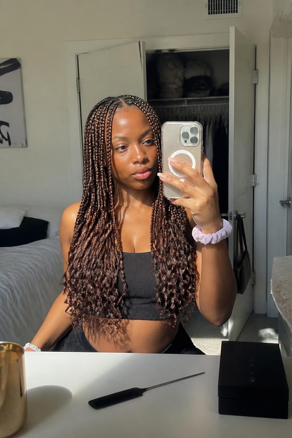 Image prompt: Photo of a Black woman wearing long chestnut knotless braids with loose curled ends and a neat middle part, candid mirror selfie, sun-drenched bedroom with a slightly open closet behind her, natural window light, containing a silk scrunchie on her wrist and a rat-tail comb on the dresser, realistic getting-ready moment, latest iPhone photo quality.