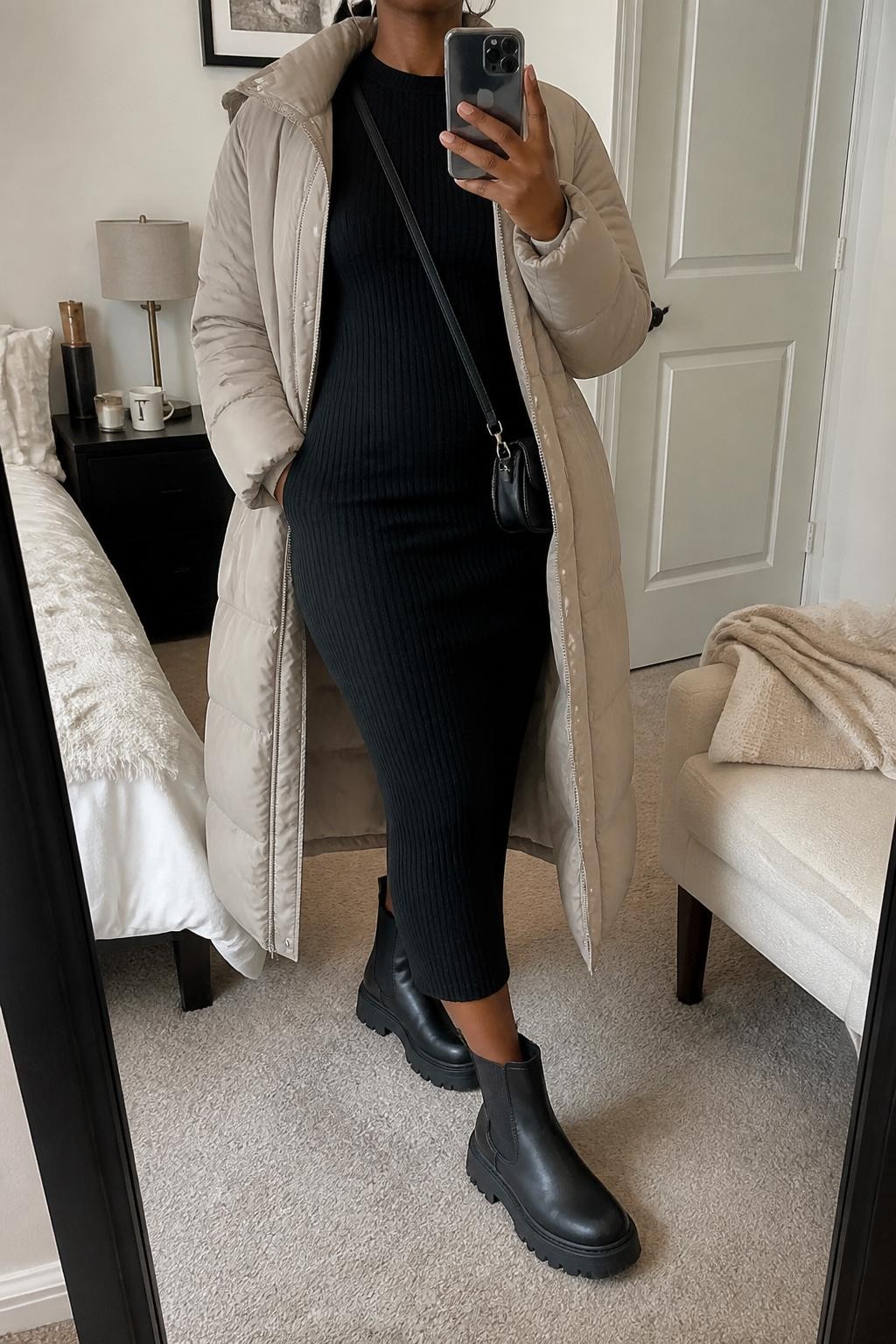 Mirror selfie of a Black woman wearing a long stone beige puffer coat left open over a fitted black ribbed midi knit dress, clearly showing chunky black Chelsea boots, a compact black crossbody bag, and silver hoop earrings, holding an iPhone in front of a cozy bedroom mirror setup, cropped at the neck so the face does not show, relaxed casual selfie pose, soft indoor lighting, a coffee mug on a bedside table and a folded blanket on a chair, natural unstaged setting, latest iPhone photo quality.