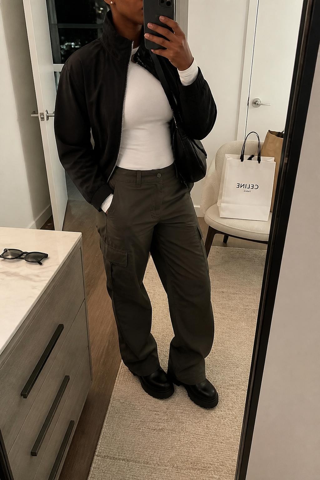mirror selfie of a Black woman wearing the exact outfit described above, clearly showing a black funnel neck jacket layered over a fitted white long-sleeve top, dark olive cargo pants, black platform sneakers, a black nylon crossbody bag, and silver stud earrings, holding a phone in front of a mirror, cropped at NECK so the face does not show, with a relaxed casual selfie pose, in a stylish city apartment, soft indoor lighting, with shopping bags beside a chair and sunglasses placed on a dresser, latest iPhone photo quality.