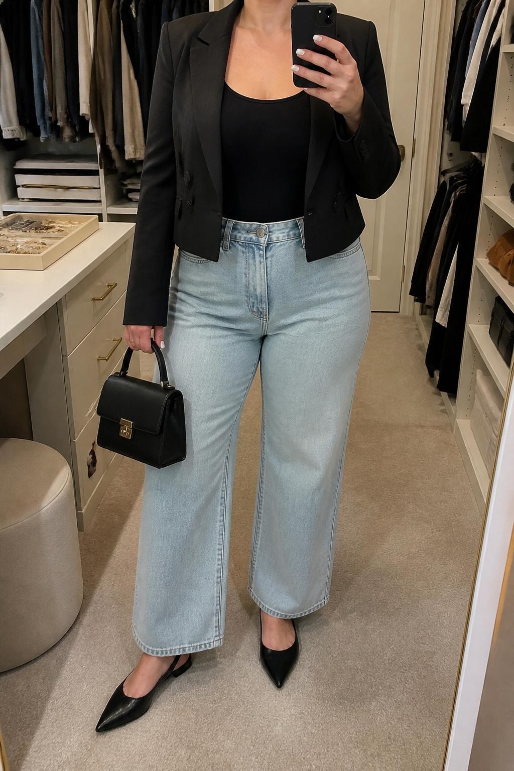 Mirror selfie of a plus size woman wearing light-wash high-waisted wide-leg jeans, a fitted black scoop-neck tank top, a structured black cropped blazer worn open, black pointed slingback flats, and a small black leather top-handle bag, clearly showing the exact clothing items, colors, denim wash, blazer shape, footwear, and accessories listed, holding a phone in front of a mirror, cropped at the neck so her face does not appear, relaxed polished selfie stance, in a walk-in closet mirror with soft indoor lighting, an open closet with clothes visible and a jewelry tray on the vanity, realistic smart-casual styling, latest iPhone photo quality.