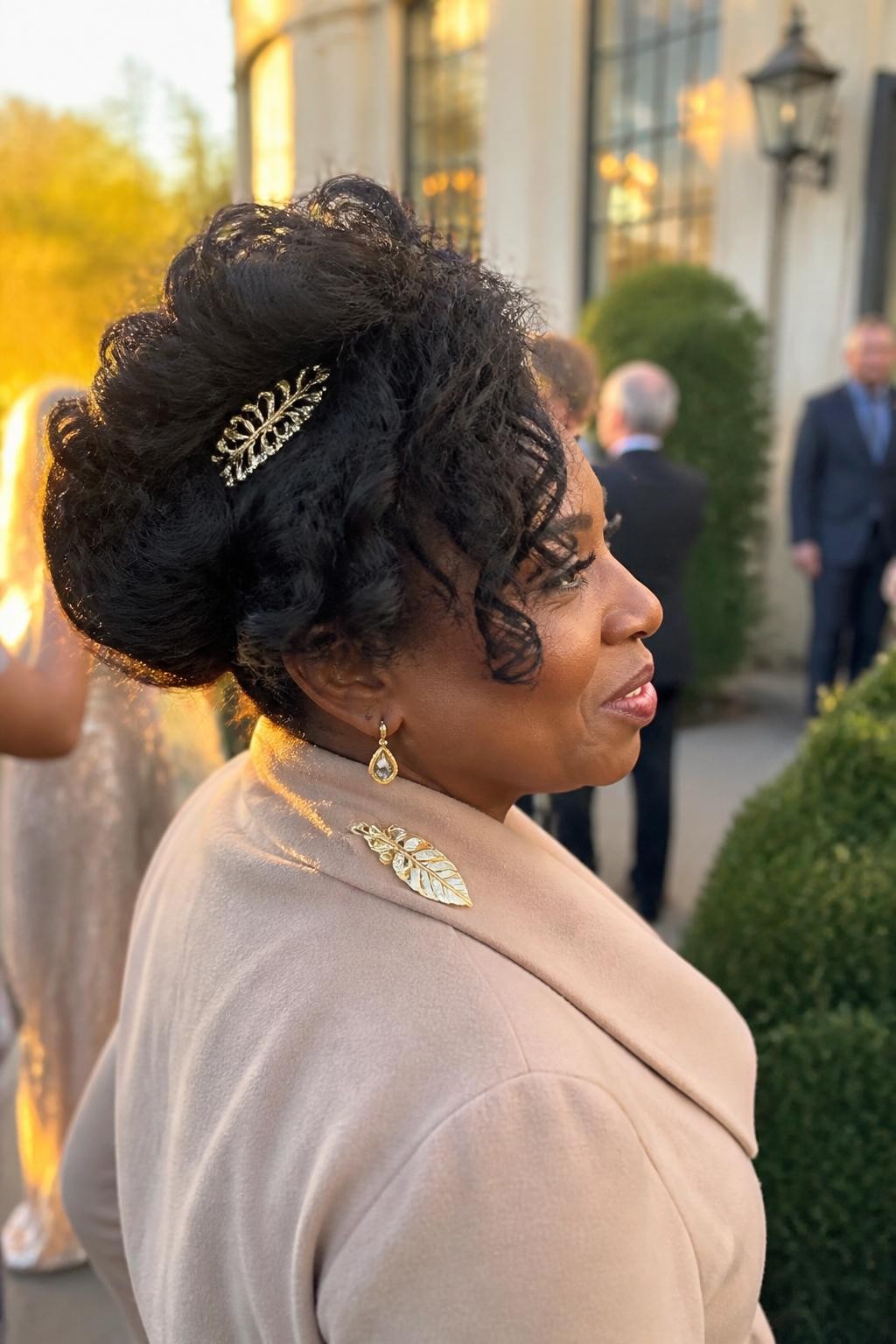 Image prompt: Photo of a Black woman wearing a full curly side-swept updo gathered above one shoulder with a small gold decorative comb, side profile view, softly blurred city wedding venue background, golden hour glow, containing drop earrings peeking through and a hair clip attached to her coat collar, realistic candid fall wedding moment, latest iPhone photo quality.