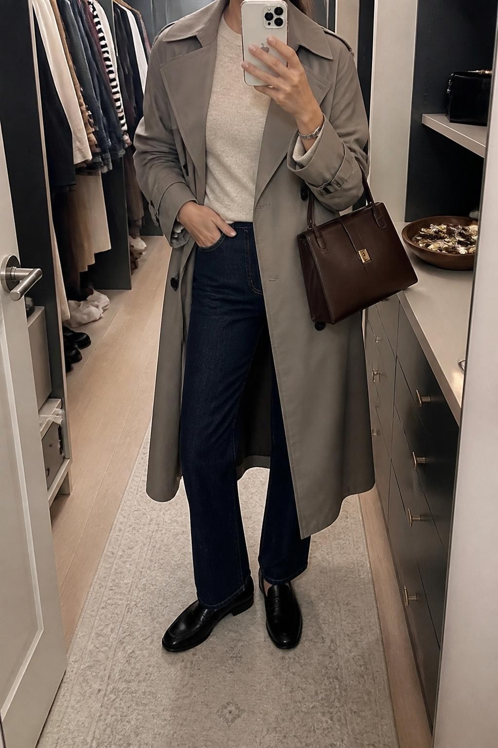 Mirror selfie of a woman wearing a relaxed taupe trench coat over an oatmeal crew-neck sweater and dark indigo straight-leg jeans, clearly showing black leather loafers, a dark brown rectangular handbag, and a slim silver wristwatch, holding an iPhone in front of a walk-in closet mirror, cropped at the neck so the face does not show, relaxed casual selfie pose, soft indoor lighting, an open closet with clothes visible and a jewelry tray on the vanity, natural unstaged setting, latest iPhone photo quality.