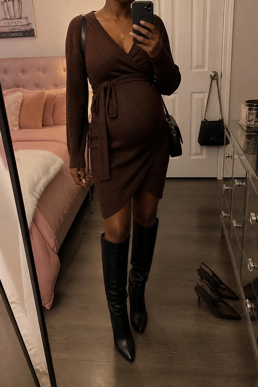 mirror selfie of a pregnant Black woman wearing a chocolate brown wrap sweater maternity dress, black knee-high leather boots, a black wool fedora, a gold pendant necklace, and a small black shoulder bag, clearly showing the same clothing items, colors, fabrics, and accessories listed in the Outfit Pieces section, holding a phone in front of a mirror, cropped at NECK so the face does not show, with a relaxed casual selfie pose, in a feminine-themed bedroom, warm moody lighting, including a pair of heels on the floor nearby and a handbag hanging on a door handle, latest iPhone photo quality.