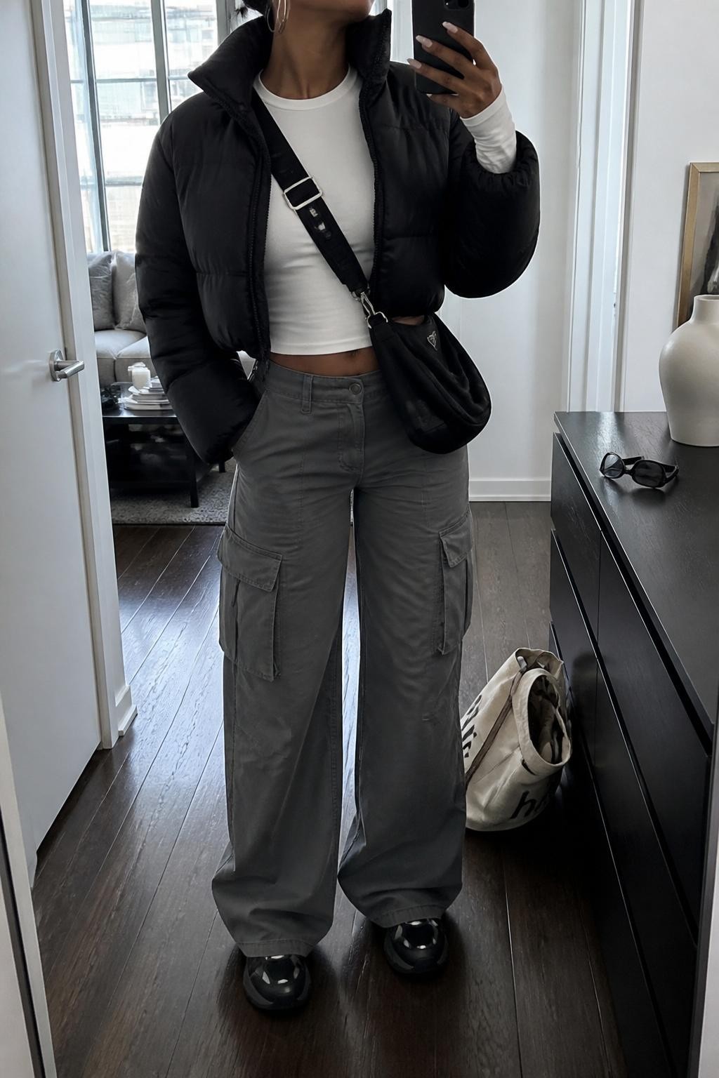 Mirror selfie of a Black woman wearing a cropped black puffer jacket, fitted white long-sleeve top, gray wide-leg cargo pants, black chunky sneakers, silver hoop earrings, and a black nylon crossbody bag, holding a phone in front of a mirror, cropped at NECK so the face does not show, relaxed casual selfie pose, in a stylish city apartment, natural window light, with sunglasses placed on a dresser and a tote bag resting on the floor, latest iPhone photo quality.