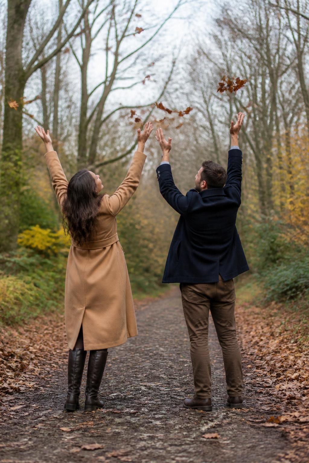 Image Prompt: A playful couple standing with their backs toward the camera on a woodland path, each holding a small handful of dry leaves and tossing them upward at the same time, the woman wearing a long camel coat and dark knee-high boots, the man wearing a navy peacoat and brown trousers, their heads turned slightly toward each other, realistic leaves caught at different heights without covering the entire scene, photographed from behind with a DSLR 35mm lens and fast shutter speed, cloudy natural light, detailed path texture, imperfect leaf colors, believable body movement, no giant leaf explosion, no jumping pose.