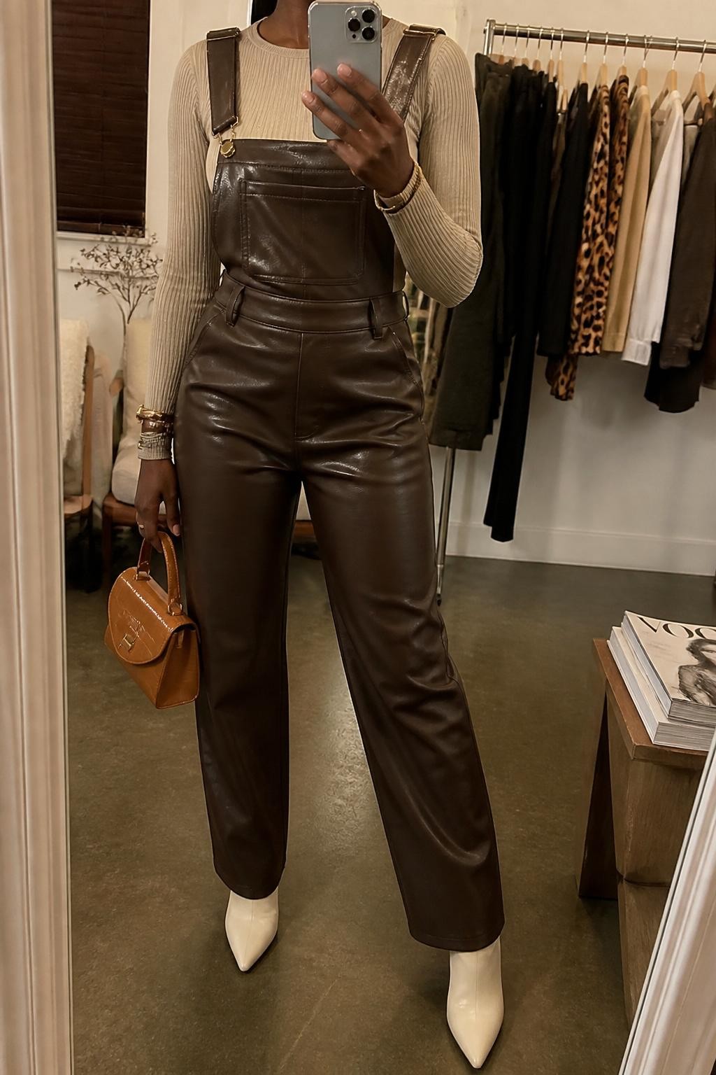 Mirror selfie of a Black woman wearing chocolate brown faux-leather overalls with a straight-leg fit over a beige long-sleeve ribbed bodysuit, clearly showing cream pointed ankle boots, a small caramel top-handle bag, and slim gold bangles, holding an iPhone in front of a fashion studio mirror corner, cropped at the neck so the face does not show, relaxed casual selfie pose, warm moody lighting, a clothing rack with garments behind and a stack of fashion magazines on a side table, natural unstaged setting, latest iPhone photo quality.