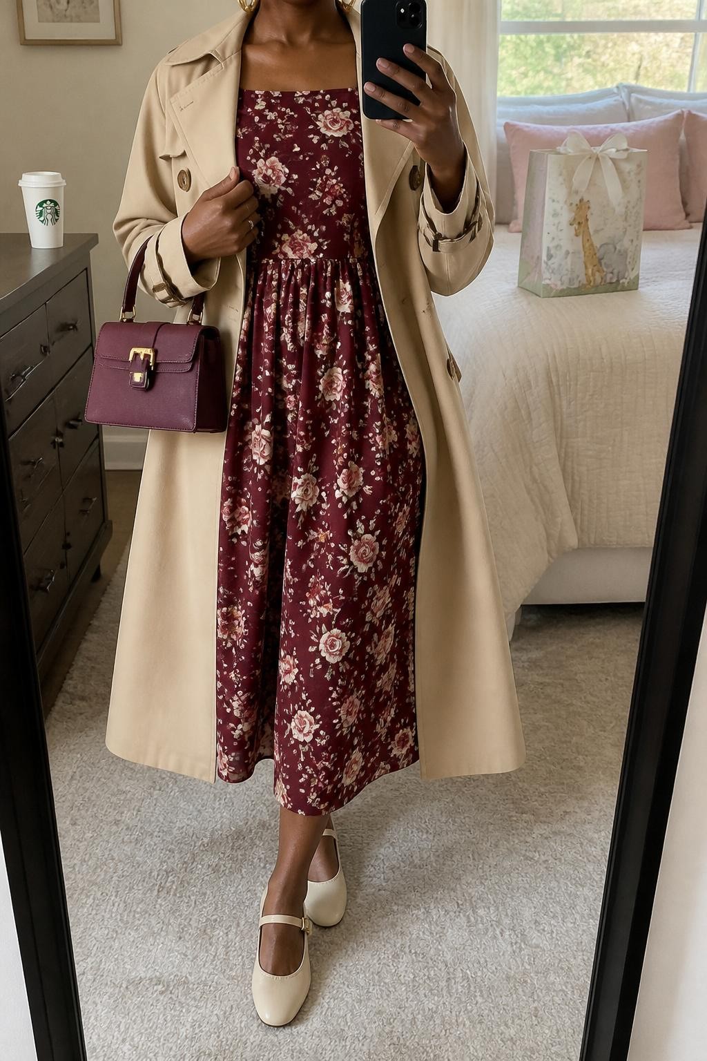 mirror selfie of a Black woman wearing the exact outfit described above, clearly showing a burgundy floral midi dress with cream and blush flowers, cream Mary Jane flats, a soft beige trench coat worn open, a burgundy mini handbag, and gold huggie earrings, holding a phone in front of a mirror, cropped at NECK so the face does not show, with a relaxed casual selfie pose, in a stylish bedroom mirror setup, natural window light, with a wrapped baby shower present on the bed and a coffee cup on the dresser, latest iPhone photo quality.