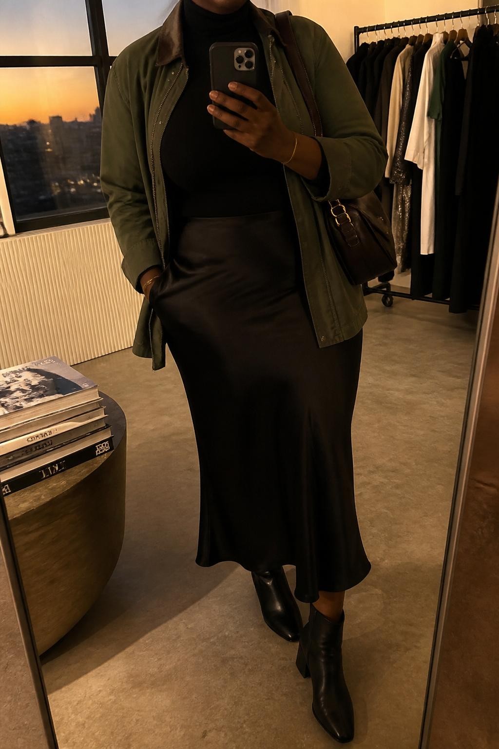 Mirror selfie of a curvy Black woman wearing an olive utility jacket, fitted black turtleneck, black satin midi slip skirt, black ankle boots, and a dark brown shoulder bag, holding a phone in front of a mirror, cropped at NECK so the face does not show, relaxed casual selfie pose, in a fashion studio mirror corner, evening golden hour, with a clothing rack with garments behind and a stack of fashion magazines on a side table, latest iPhone photo quality.