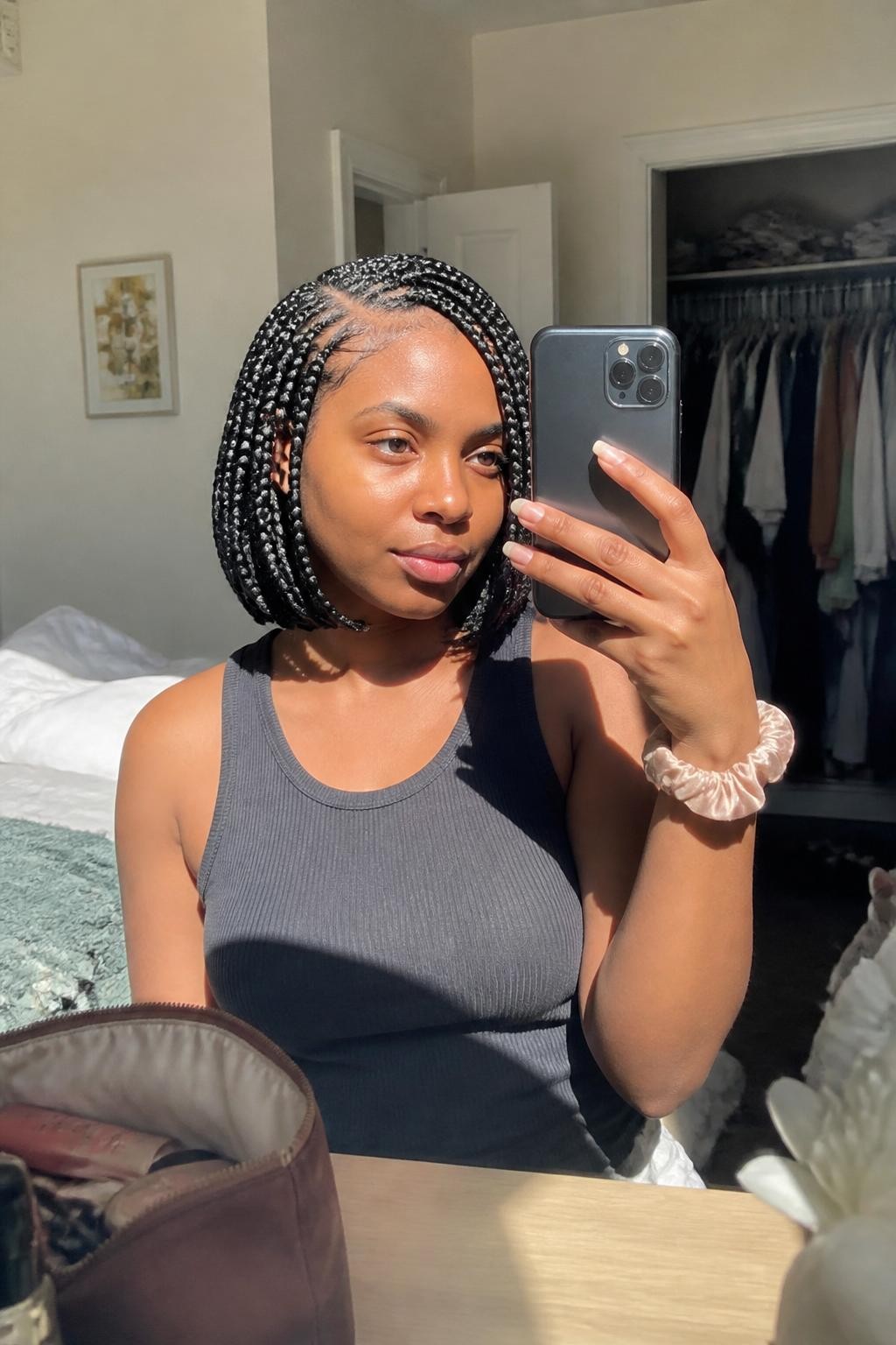 Image prompt: Photo of a Black woman wearing a chin-length box-braid bob with a deep side part and softly curved ends, candid mirror selfie, sun-drenched bedroom with a slightly open closet behind her, natural window light, containing a makeup bag on the dresser and a silk scrunchie on her wrist, realistic morning moment, latest iPhone photo quality.