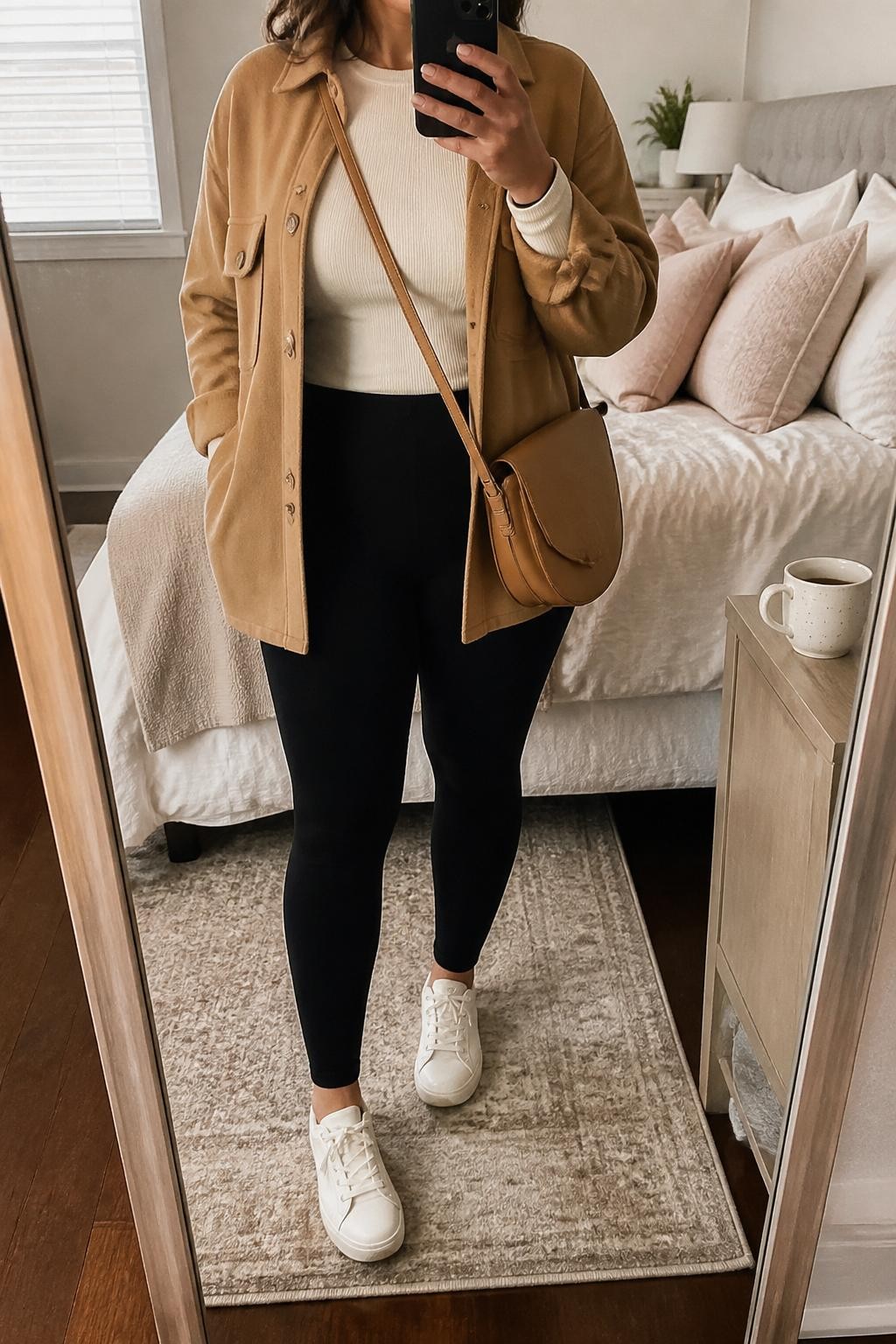Mirror selfie of a curvy woman wearing a camel oversized shacket, cream fitted long-sleeve top, black ponte leggings, white leather sneakers, and a tan crossbody bag, holding a phone in front of a mirror, cropped at NECK so the face does not show, relaxed casual selfie pose, in a cozy bedroom mirror setup, natural window light, with soft pillows on an unmade bed and a coffee mug on a bedside table, latest iPhone photo quality.