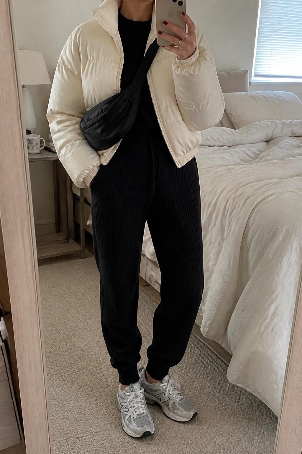 Mirror selfie of a woman wearing a black knit jogger set, short ivory puffer jacket, gray chunky sneakers, cream knit beanie, and a black nylon crossbody bag, holding a phone in front of a mirror, cropped at NECK so the face does not show, relaxed casual selfie pose, in a cozy bedroom mirror setup, soft indoor lighting, with soft pillows on an unmade bed and a coffee mug on a bedside table, latest iPhone photo quality.