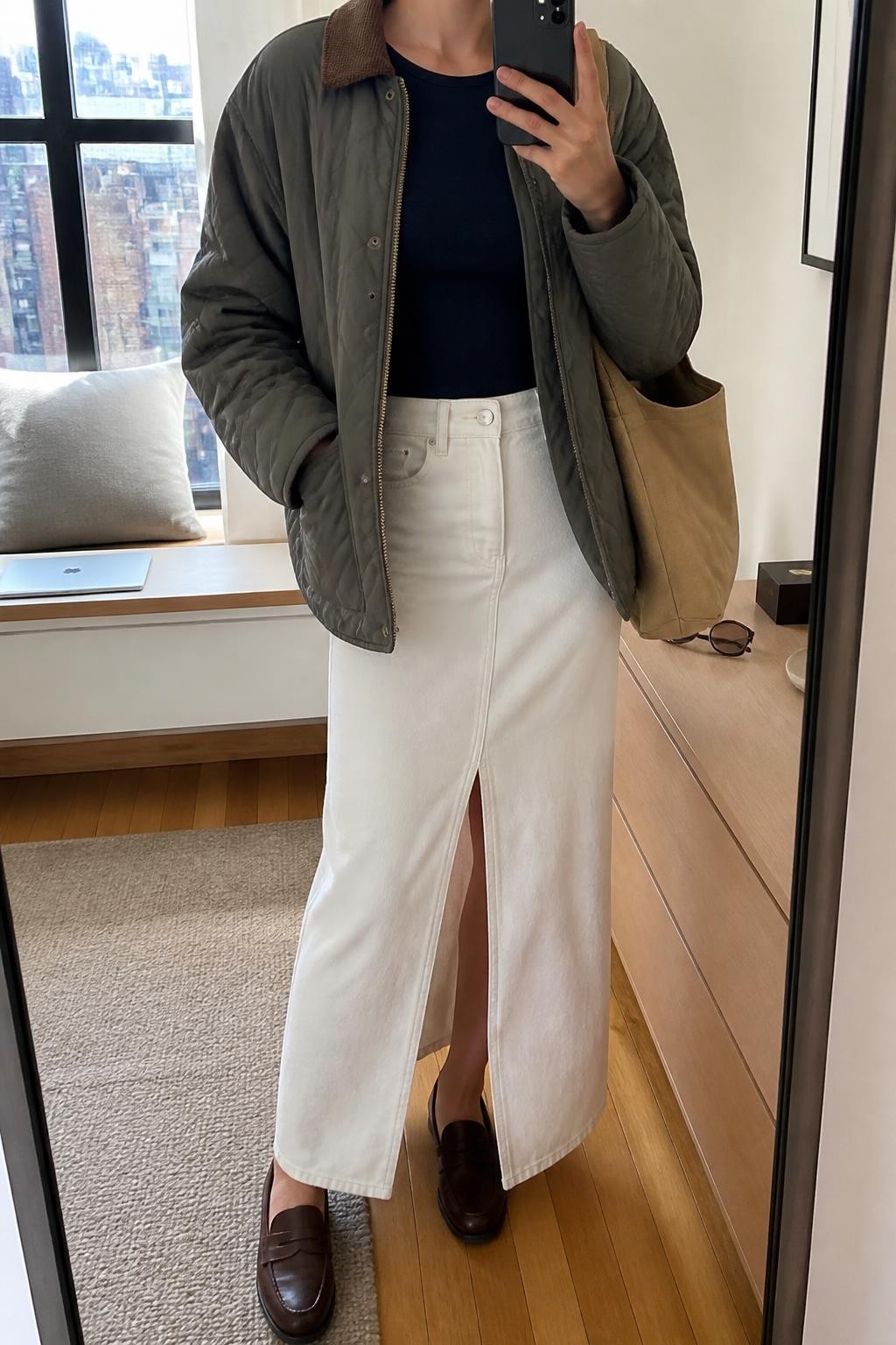 Mirror selfie of a woman wearing an olive-grey quilted jacket with a brown corduroy collar, fitted navy long-sleeve top, white denim maxi skirt with a front slit, dark brown leather loafers, and a tan canvas tote bag, clearly showing the exact clothing items, colors, fabrics, and accessories listed, holding a phone in front of a stylish city apartment mirror, cropped at the neck so the face does not show, relaxed casual selfie pose, natural window light, a laptop on a bench and sunglasses placed on a dresser, believable everyday setting, latest iPhone photo quality.
